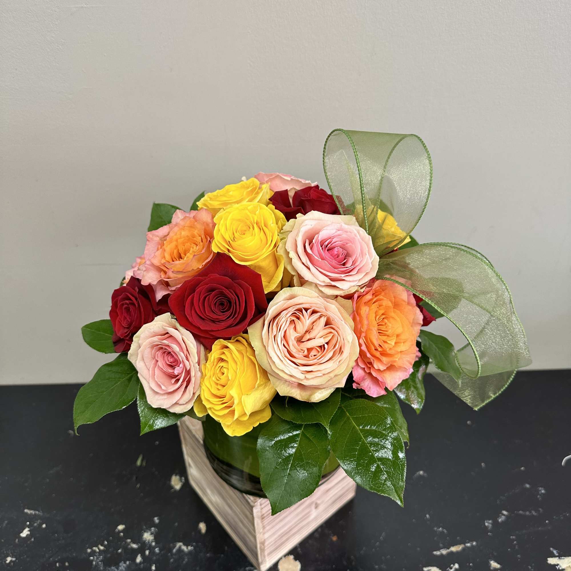 Mixed roses in pink, yellow, orange, and red in a round container with a green ribbon