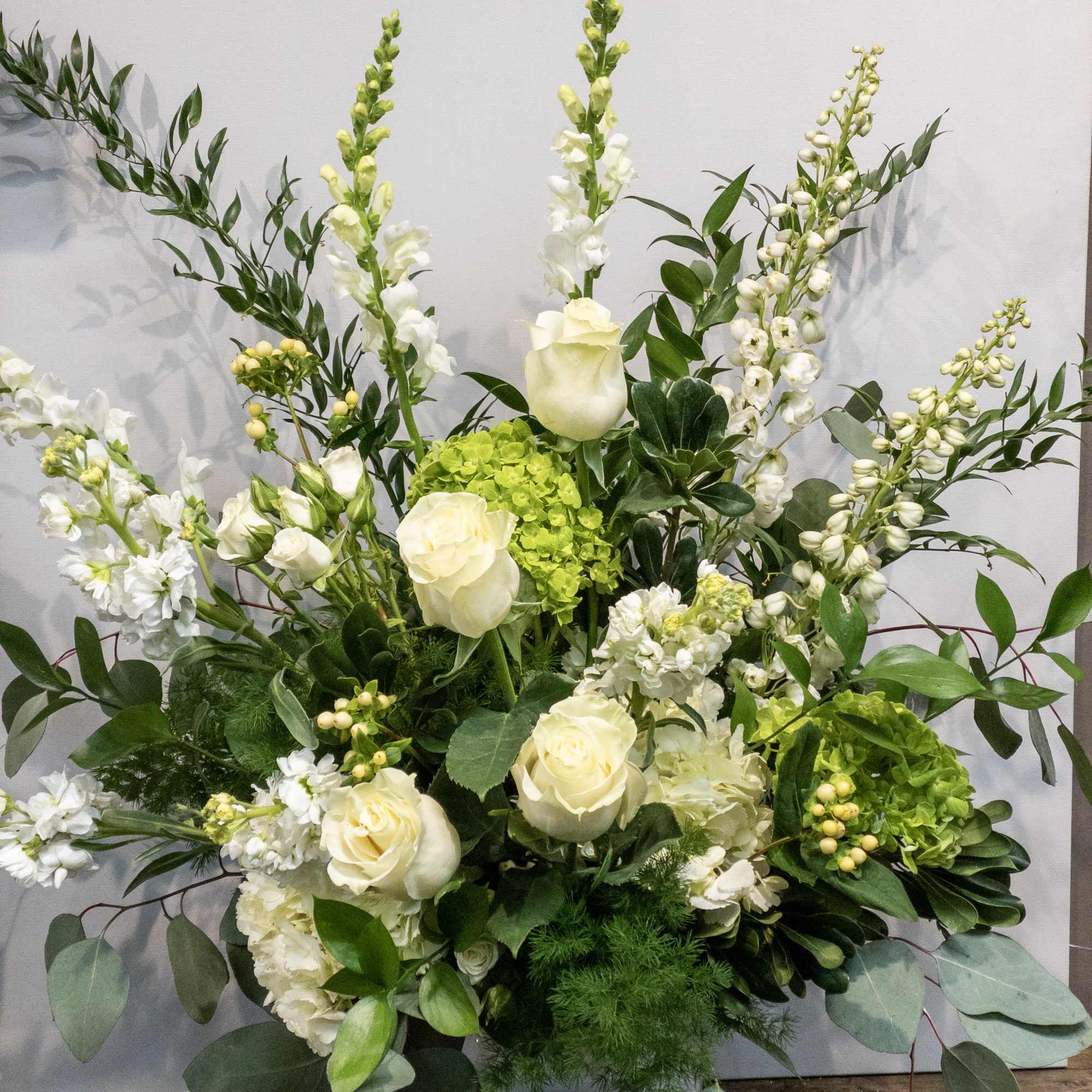 Tall white and green floral arrangement with roses, snapdragons, and hydrangeas in a white pedestal vase
