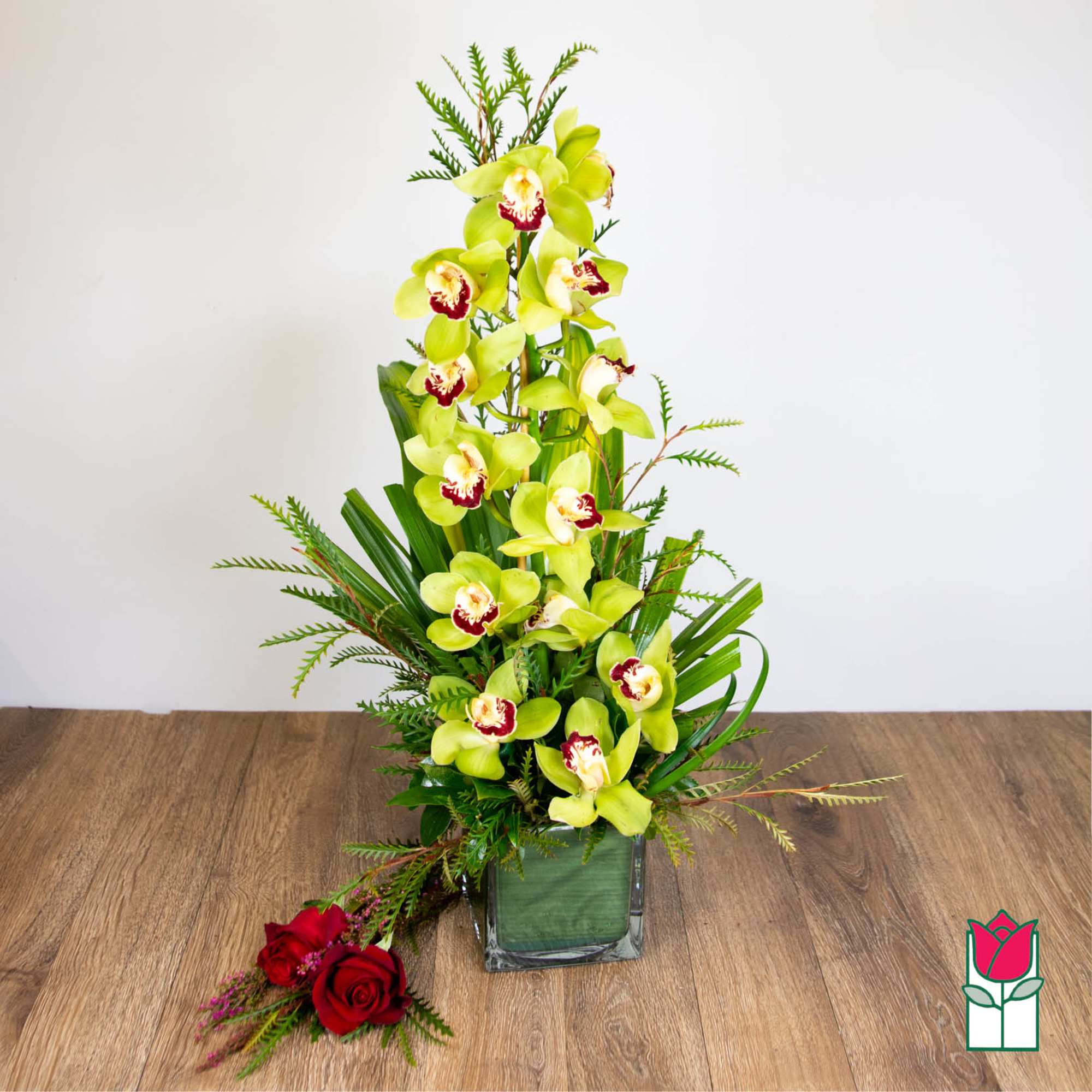 Tall green cymbidium orchid arrangement in a leaf-lined glass cube vase with two red roses beside it