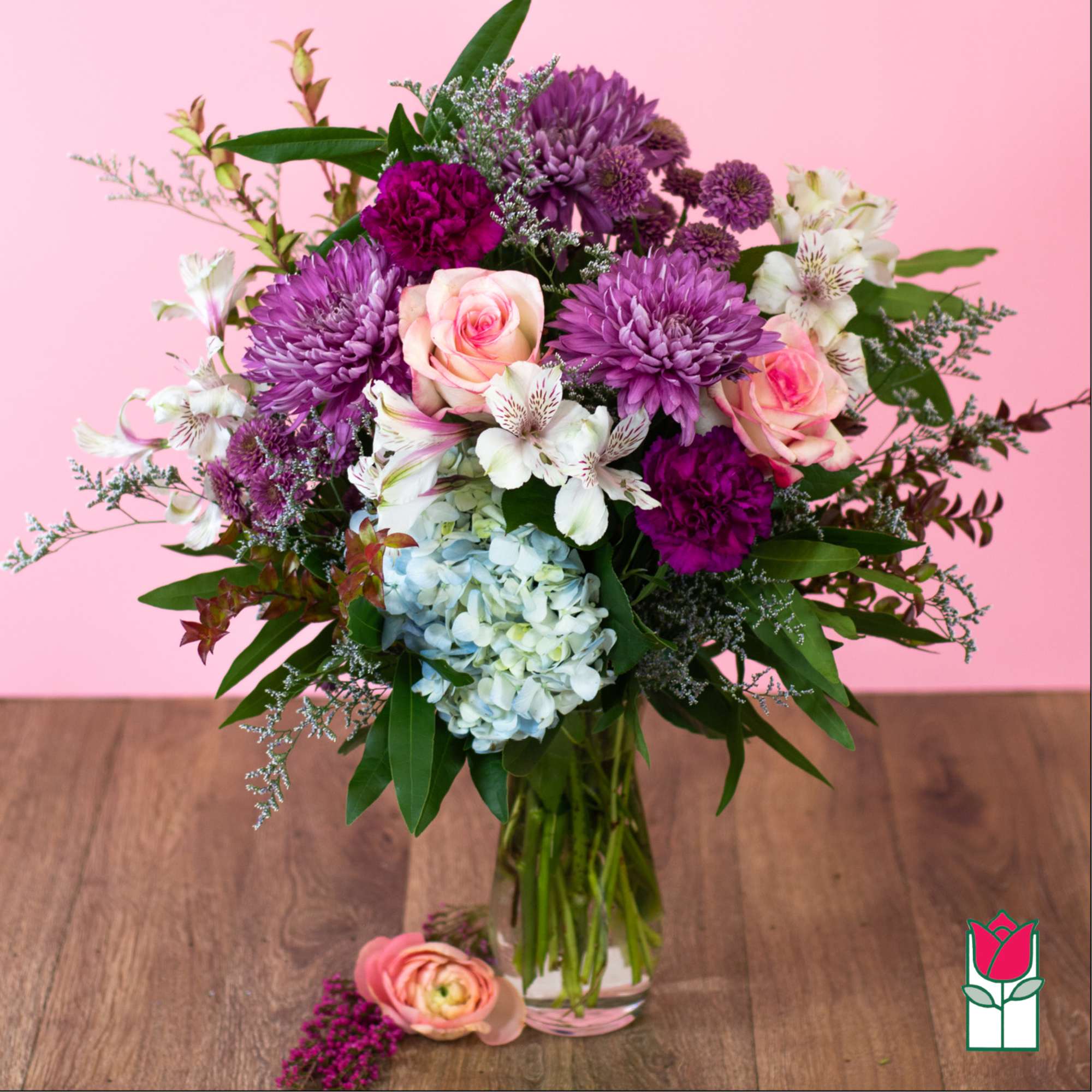 Mixed bouquet of purple mums, pink roses, white alstroemeria, and blue hydrangea in a clear glass vase.