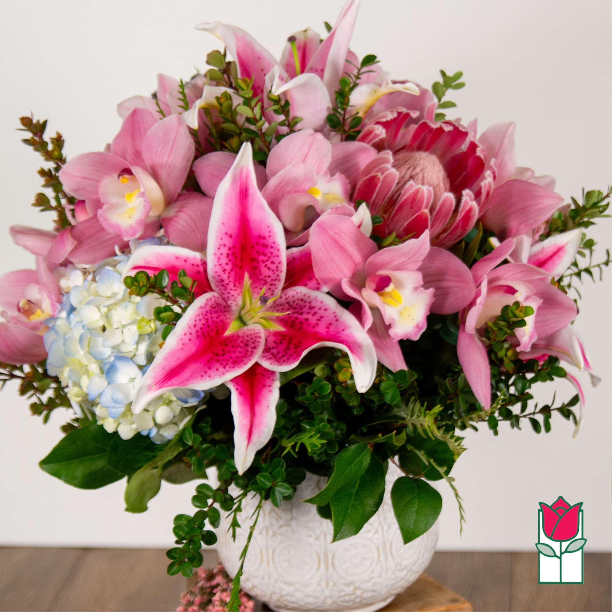 Pink lilies and orchids arranged in a white vase with blue hydrangea
