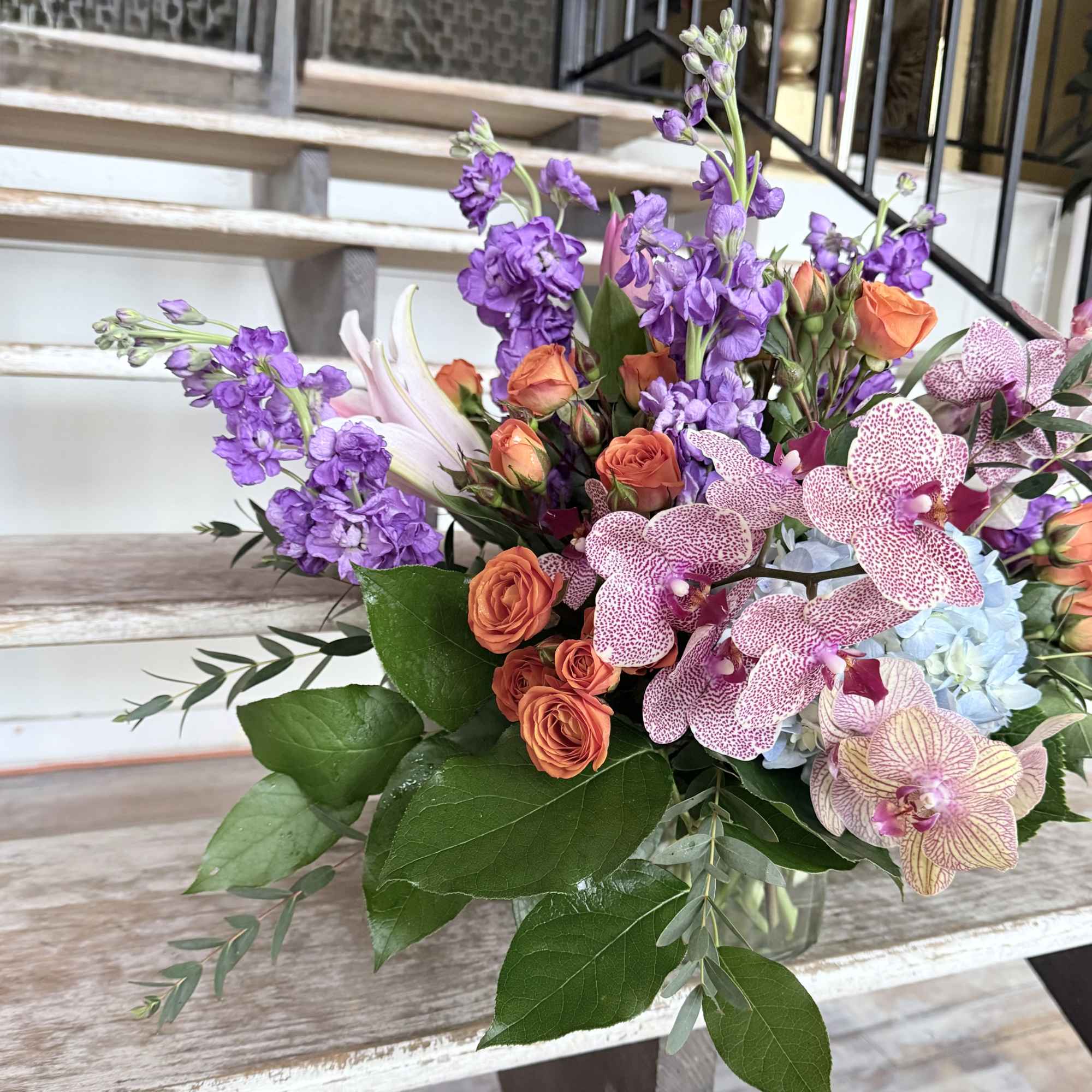 Mixed bouquet with purple stock, orange spray roses, and spotted orchids in a glass vase