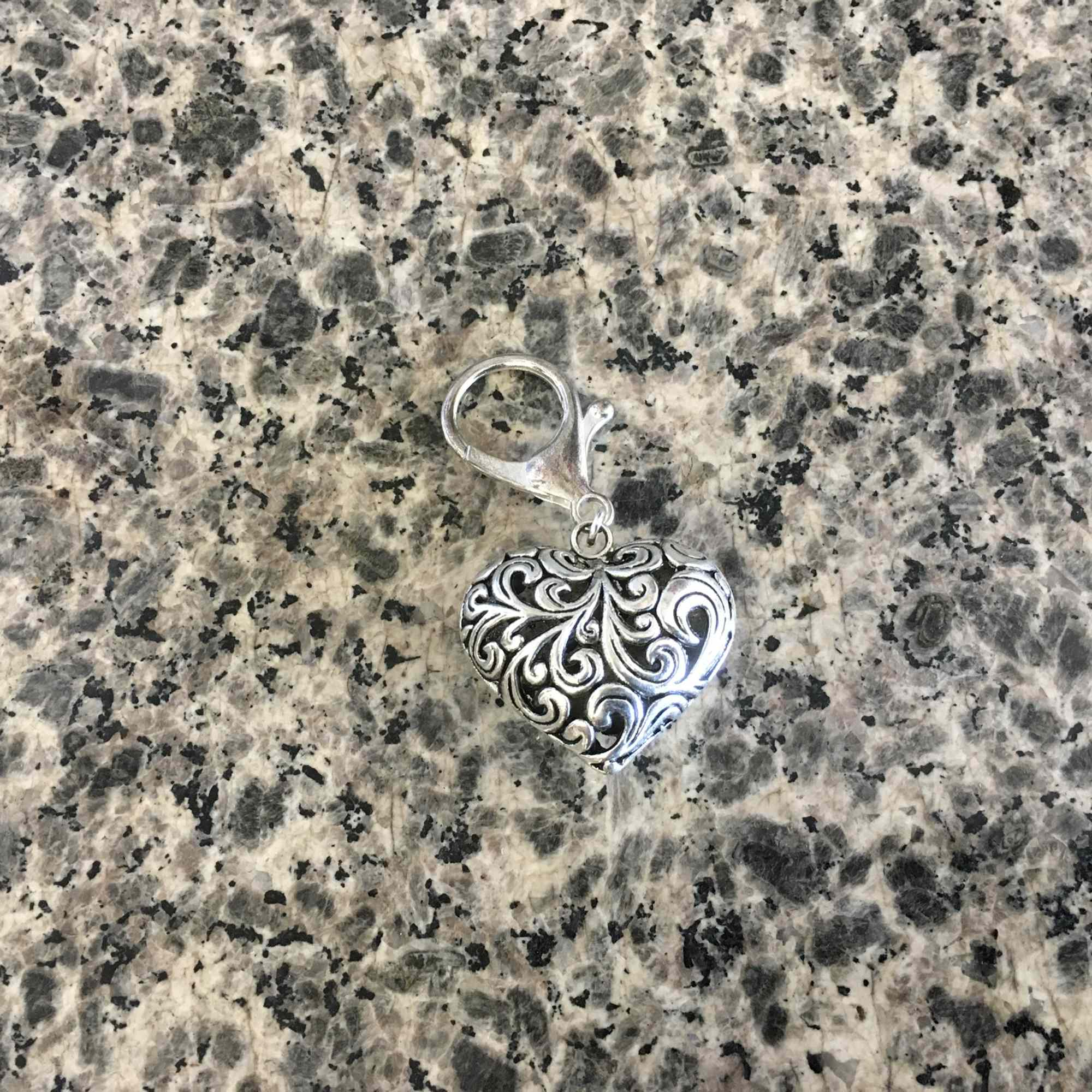 Silver heart-shaped charm with ornate scrollwork on a stone surface