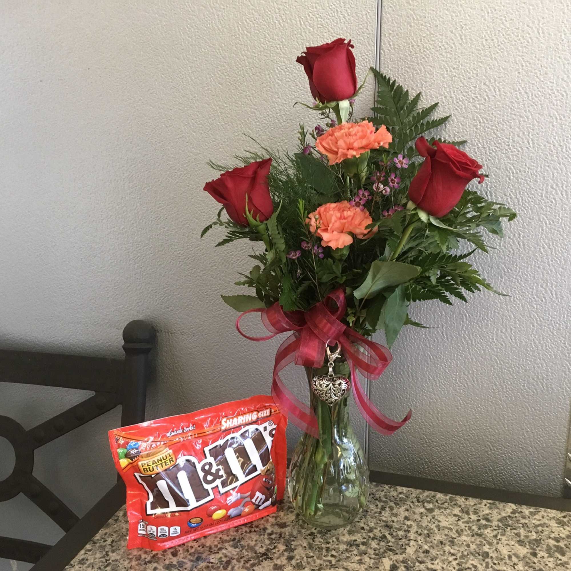 Red roses and orange carnations in a glass vase with a ribbon, beside a bag of M&M's.