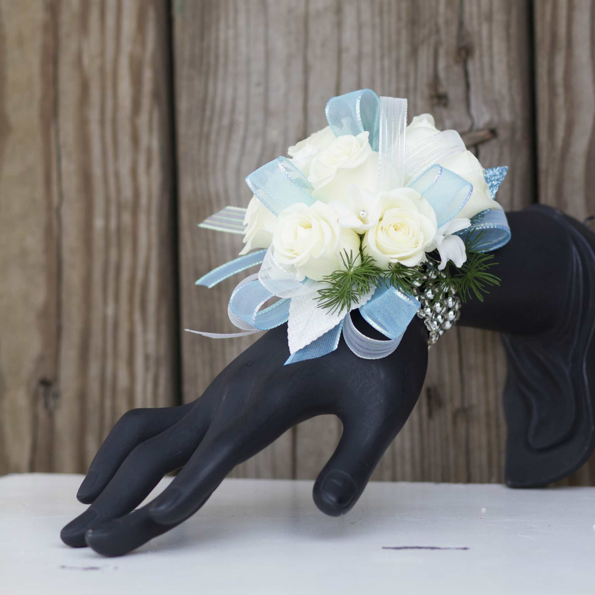 White rose wrist corsage with blue and white ribbons on jeweled band displayed on black hand stand
