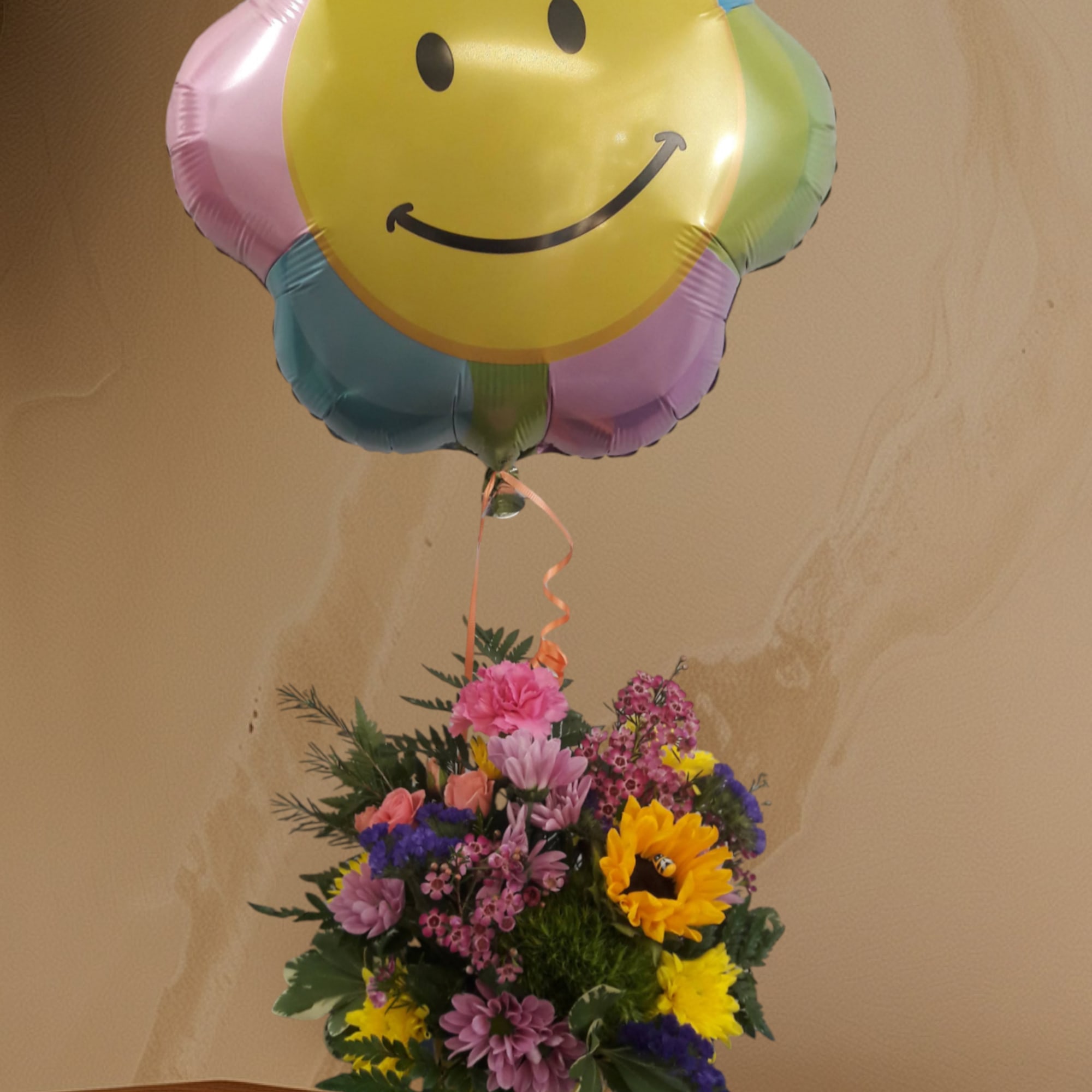Smiley-face balloon above a colorful mixed flower arrangement in a yellow container.
