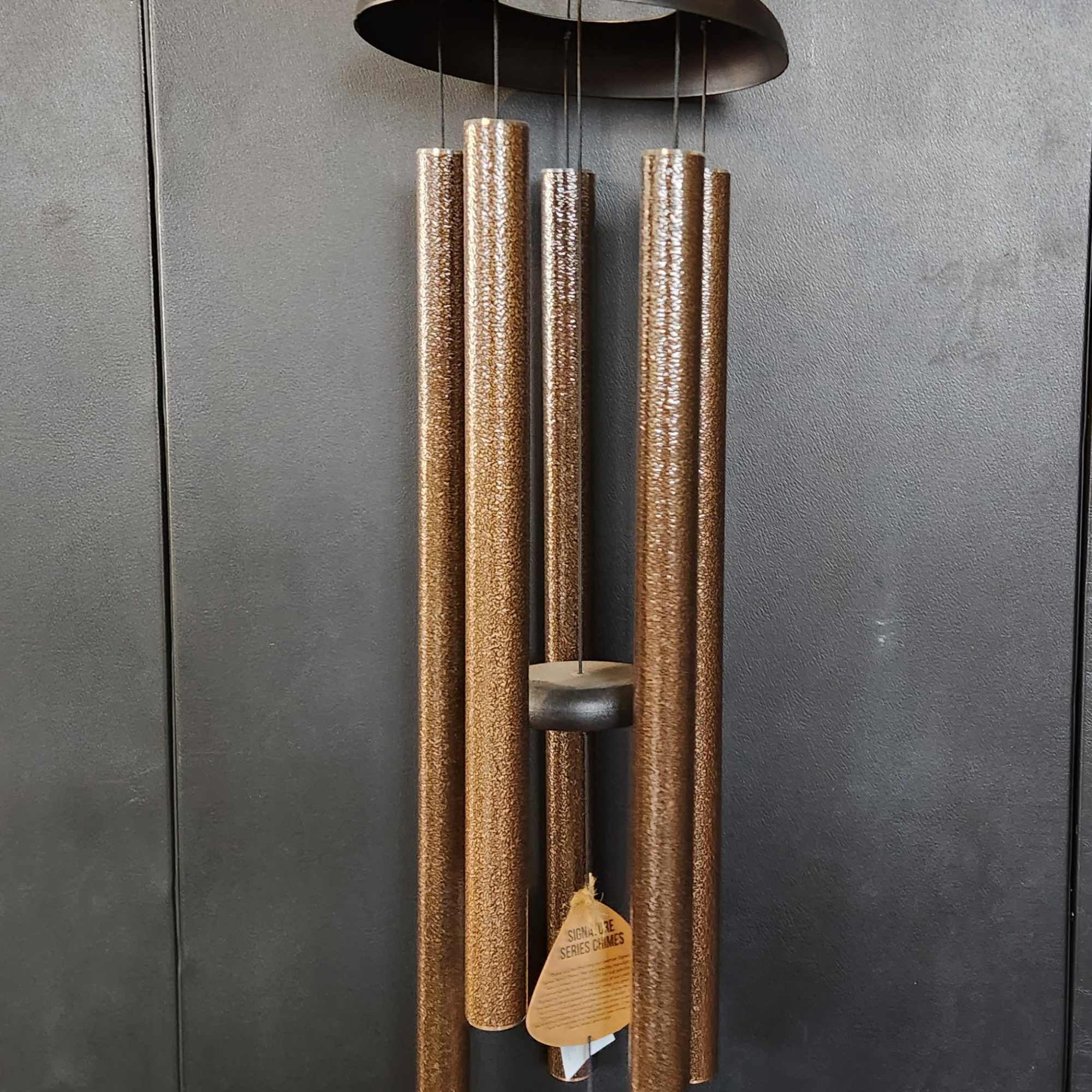 Bronze wind chime with five hanging tubes against a dark wall.