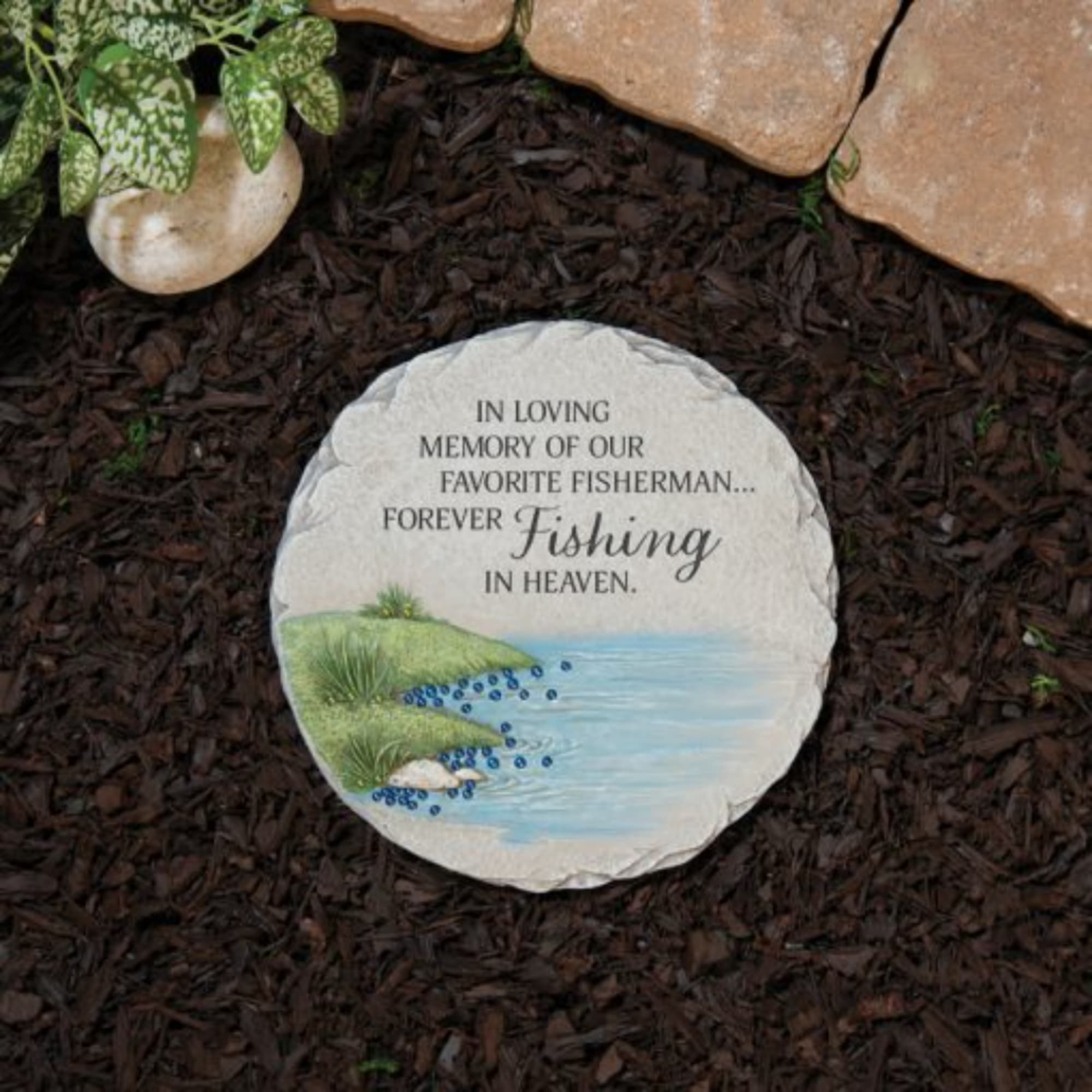 Round memorial stone with fishing tribute text in a garden bed