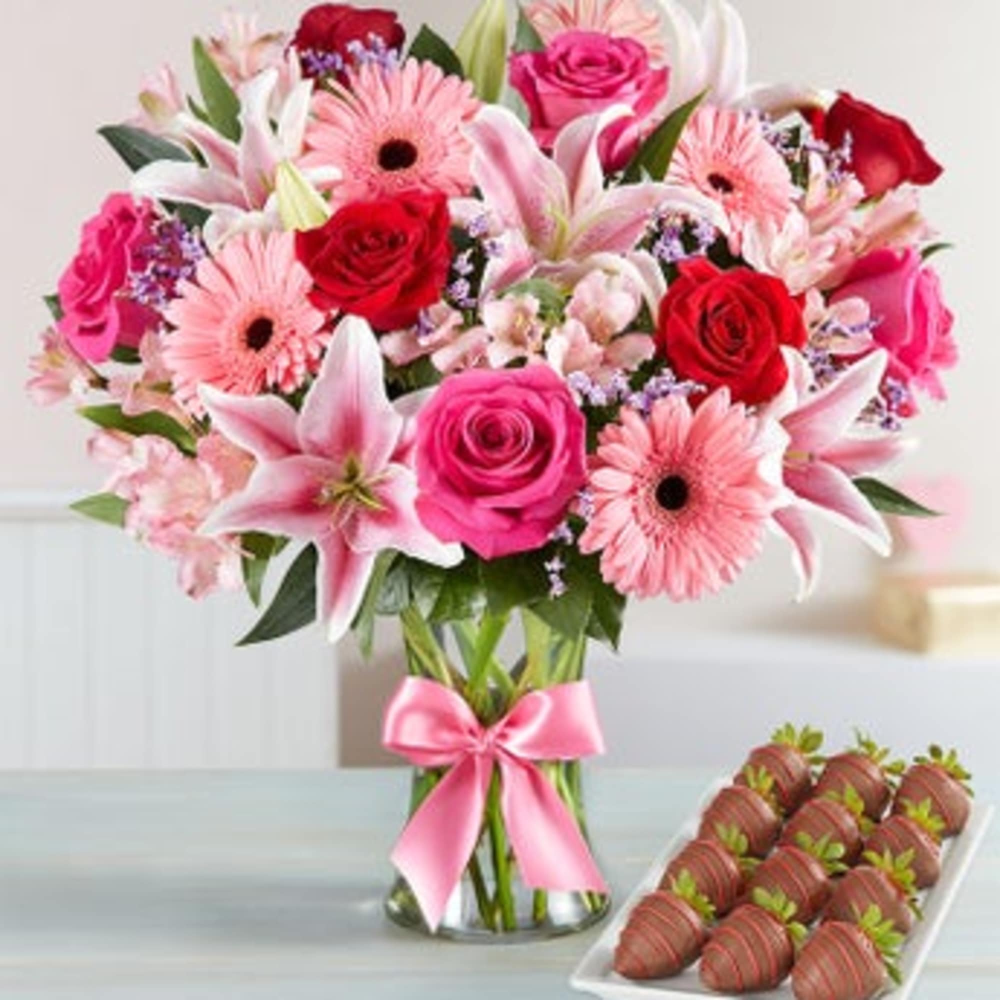 Pink and red mixed bouquet in a glass vase with a pink ribbon, beside chocolate-covered strawberries