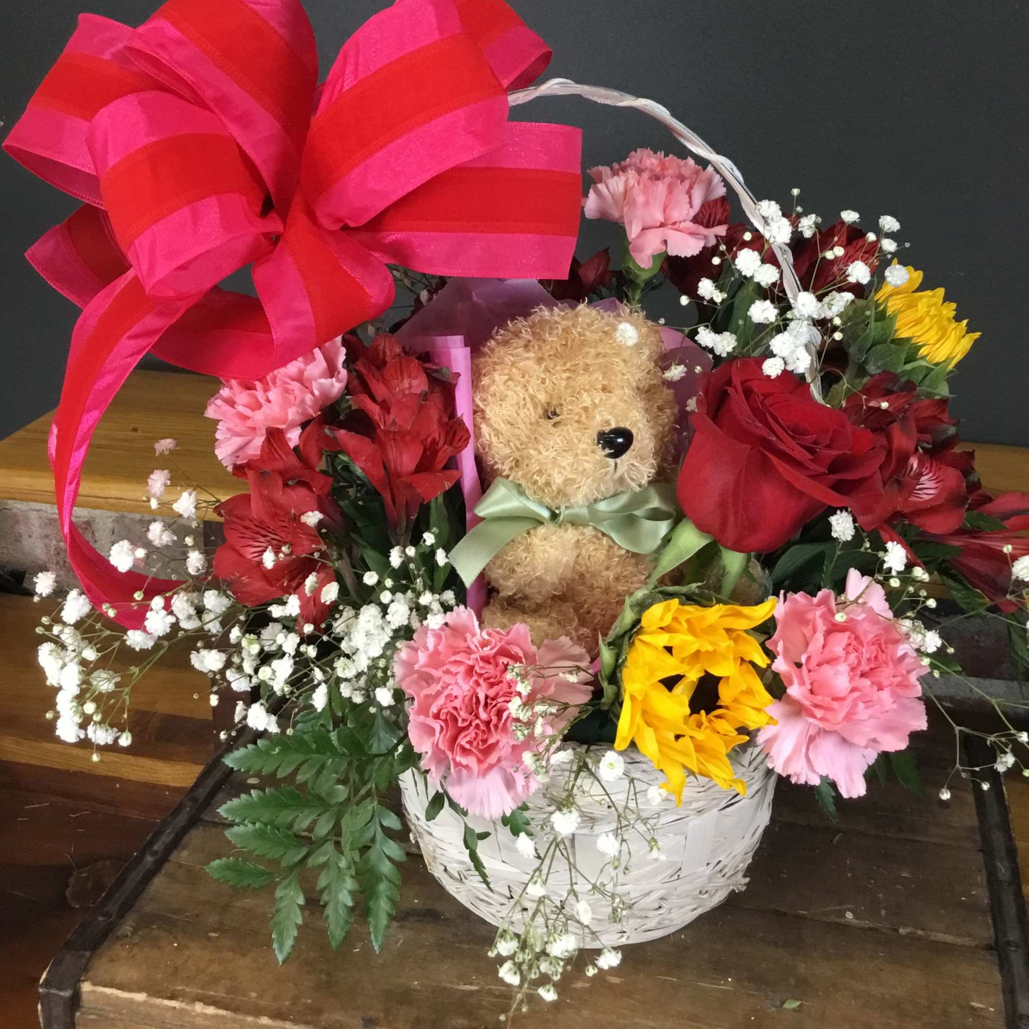 Basket arrangement with a teddy bear, roses, carnations, and a large pink bow