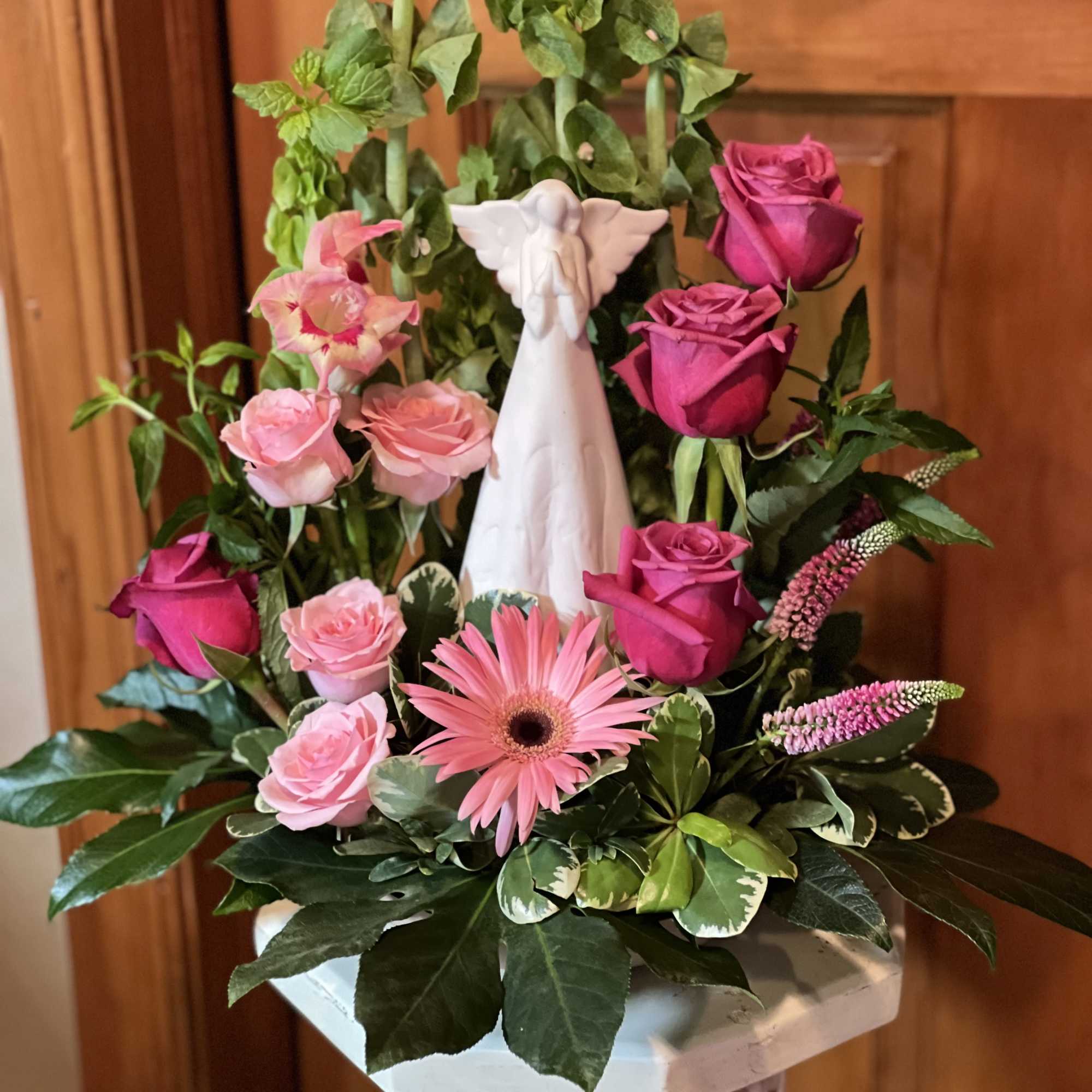Pink roses and a gerbera daisy arranged around a white angel figurine in a pedestal vase