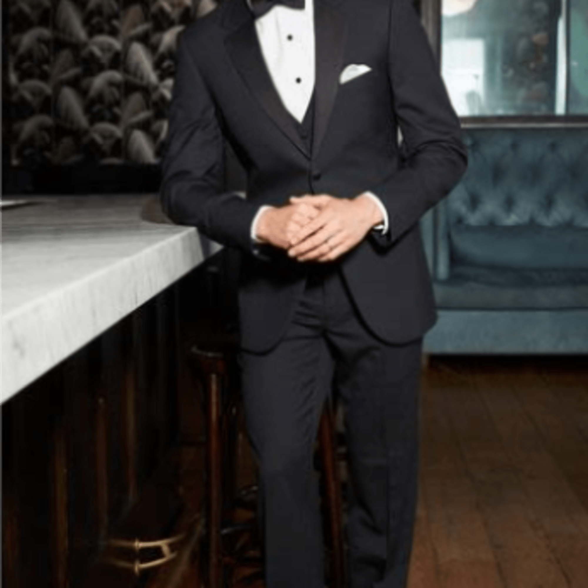 Man in a black tuxedo with bow tie standing beside a marble bar in a dim lounge