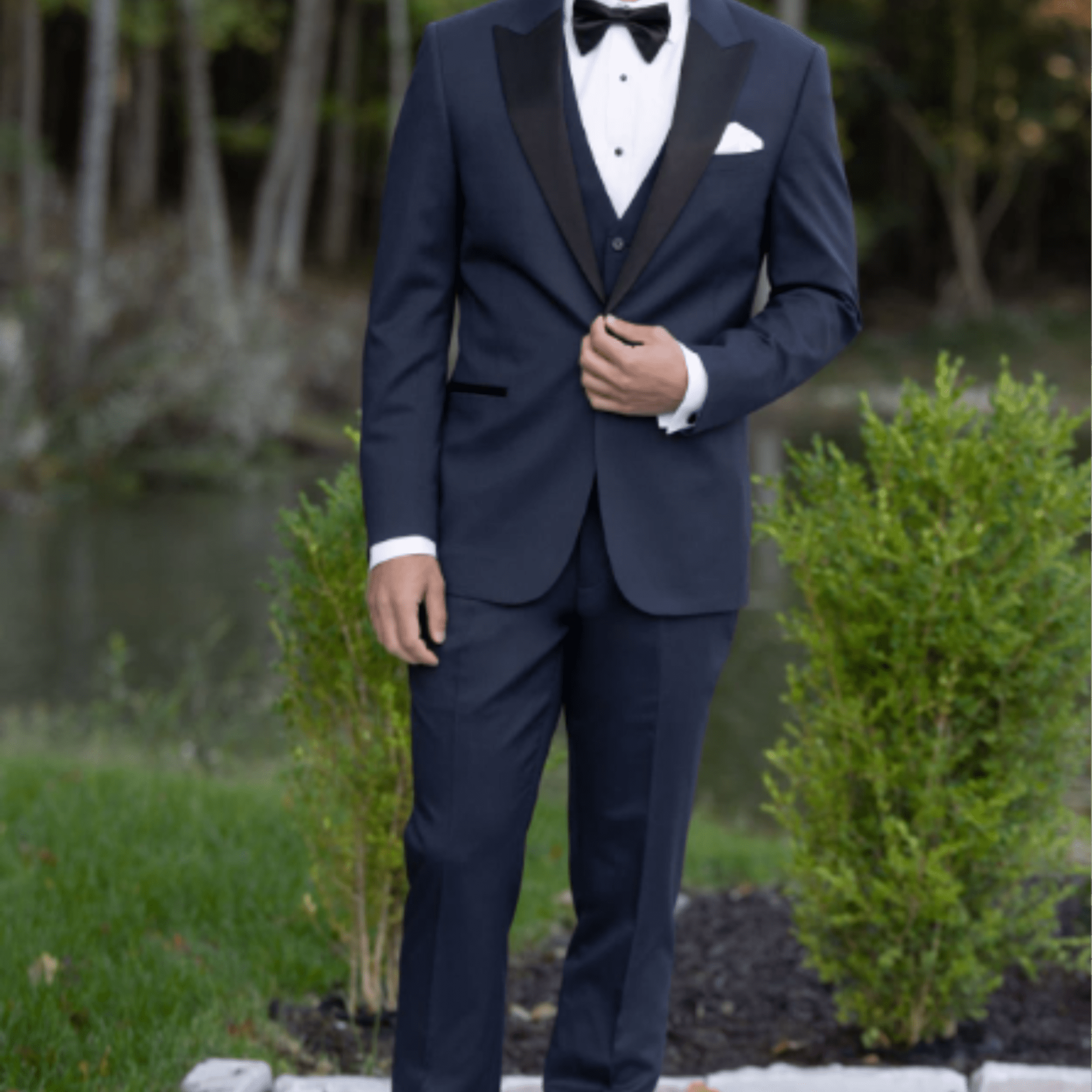 Man in a navy tuxedo with black lapels and bow tie standing on a paved path outdoors