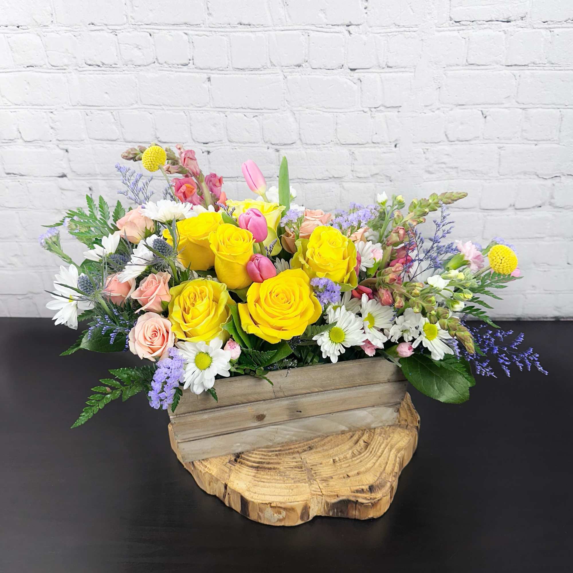 Yellow roses and pastel flowers arranged in a wooden box on a wood slice