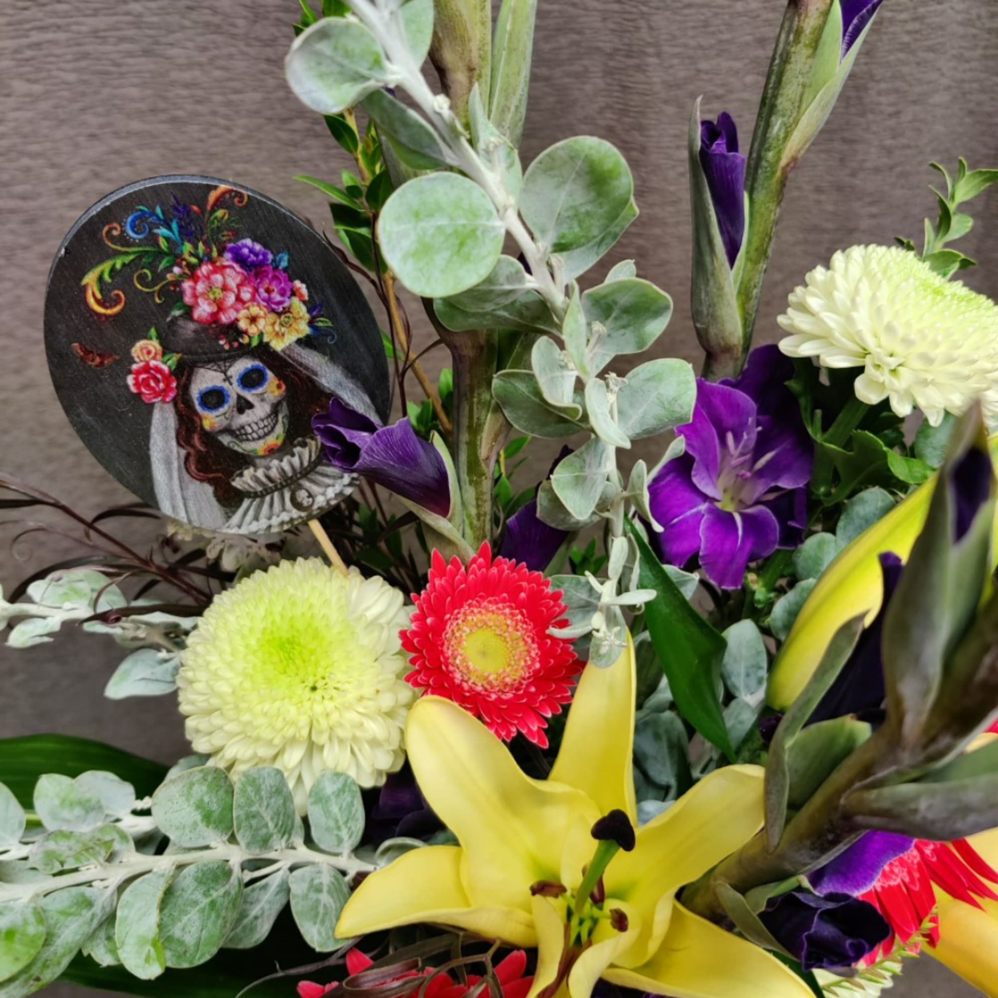 Colorful floral arrangement with yellow lilies, purple blooms, and a decorative skull ornament