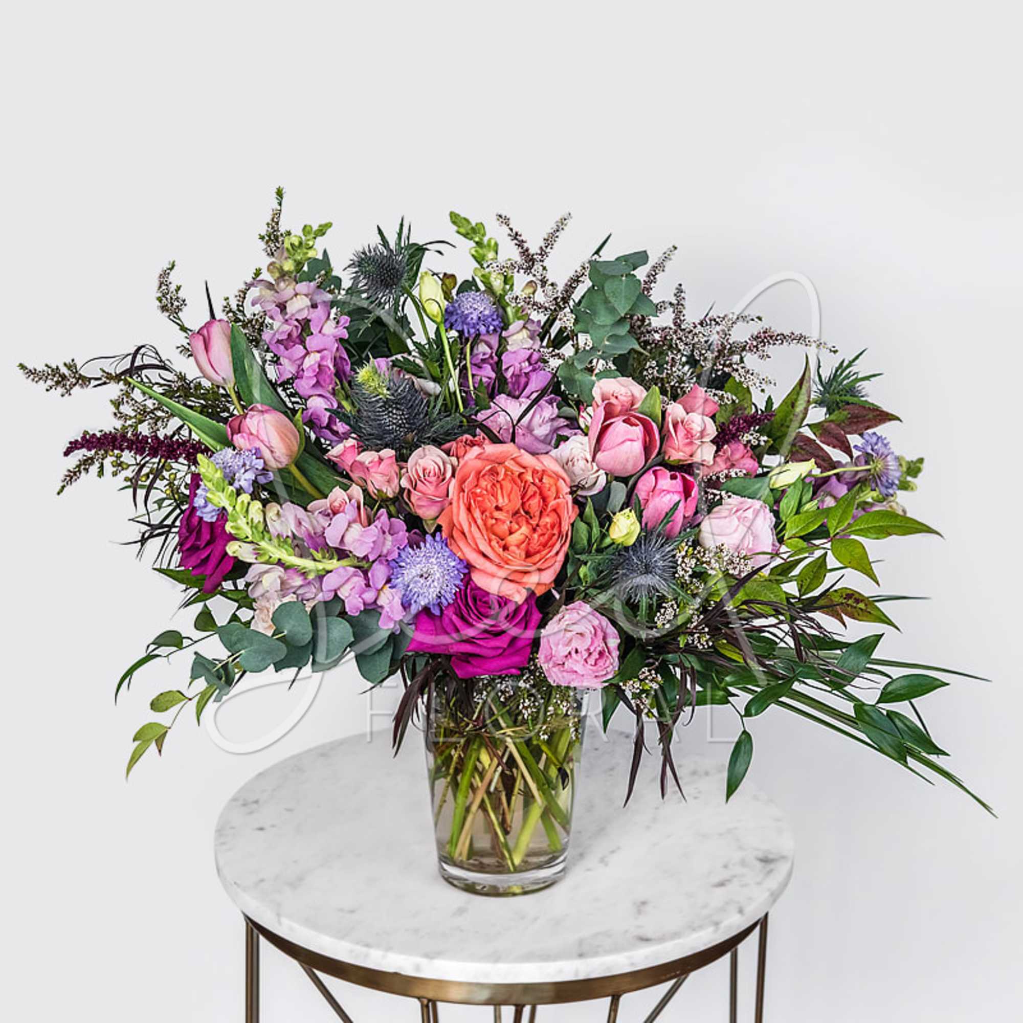 Lush arrangement of pink, purple, and peach flowers in a clear glass vase on a small round table.