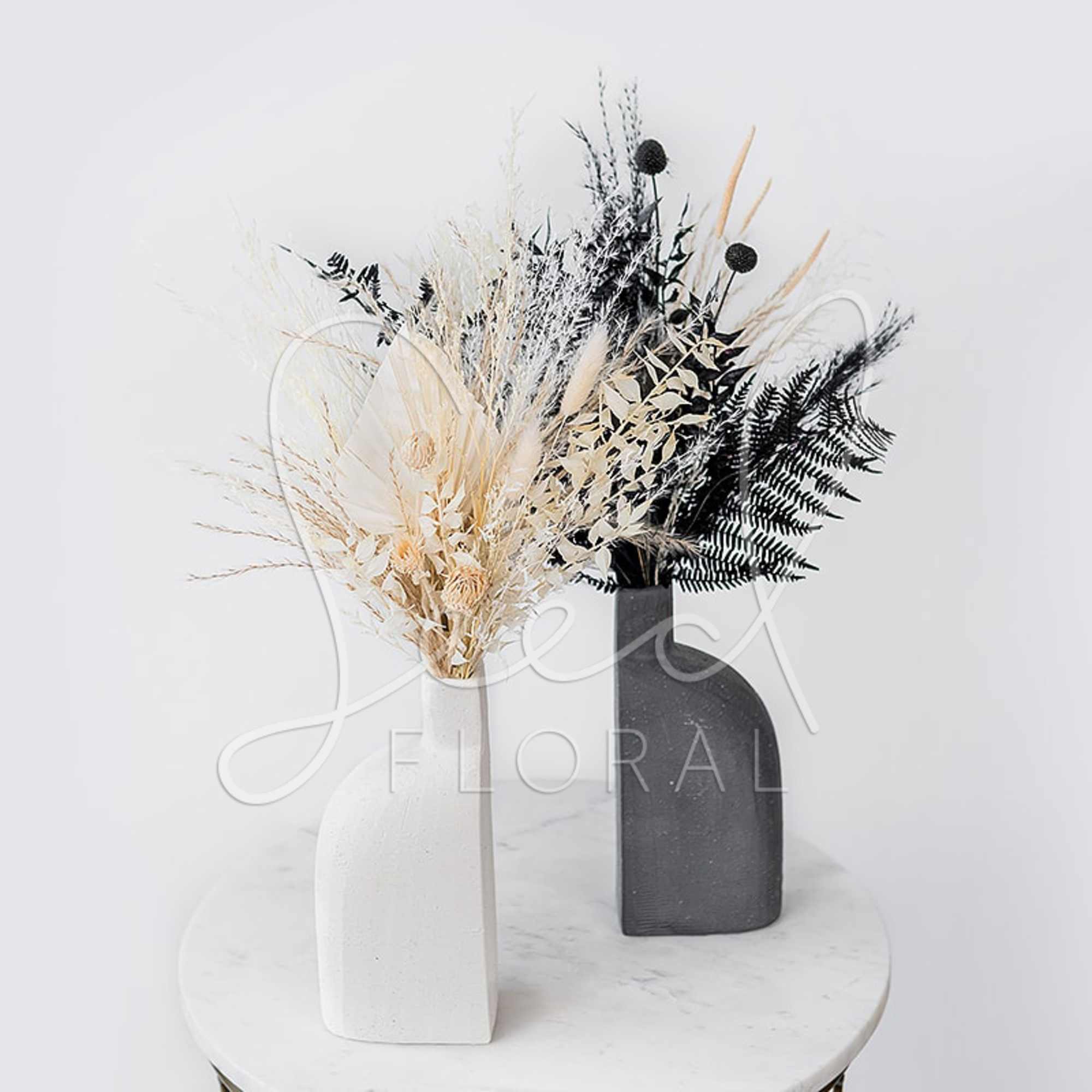 Two dried floral arrangements in white and black vases on a round table.
