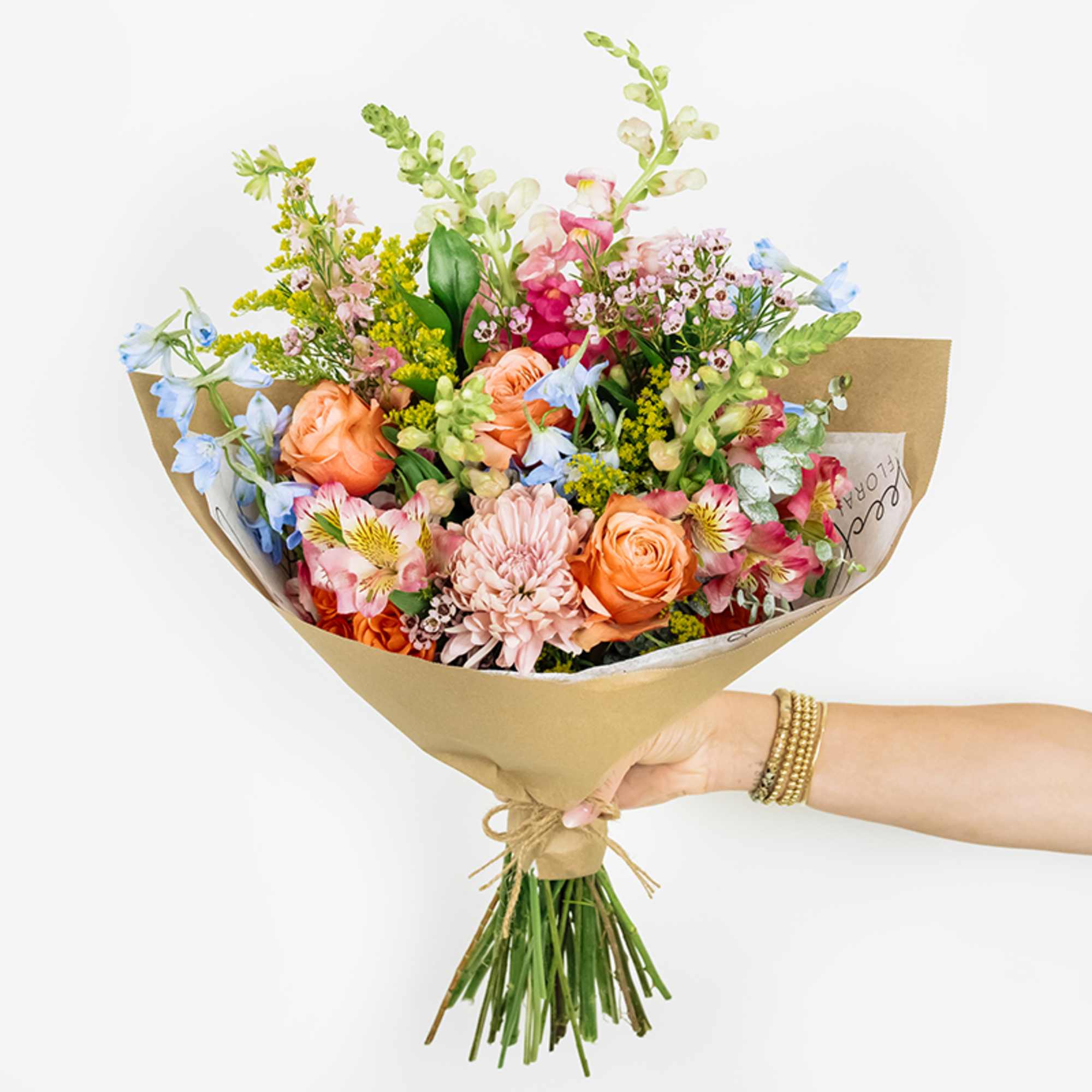 Handheld bouquet of mixed pink, peach, blue, and white flowers wrapped in kraft paper