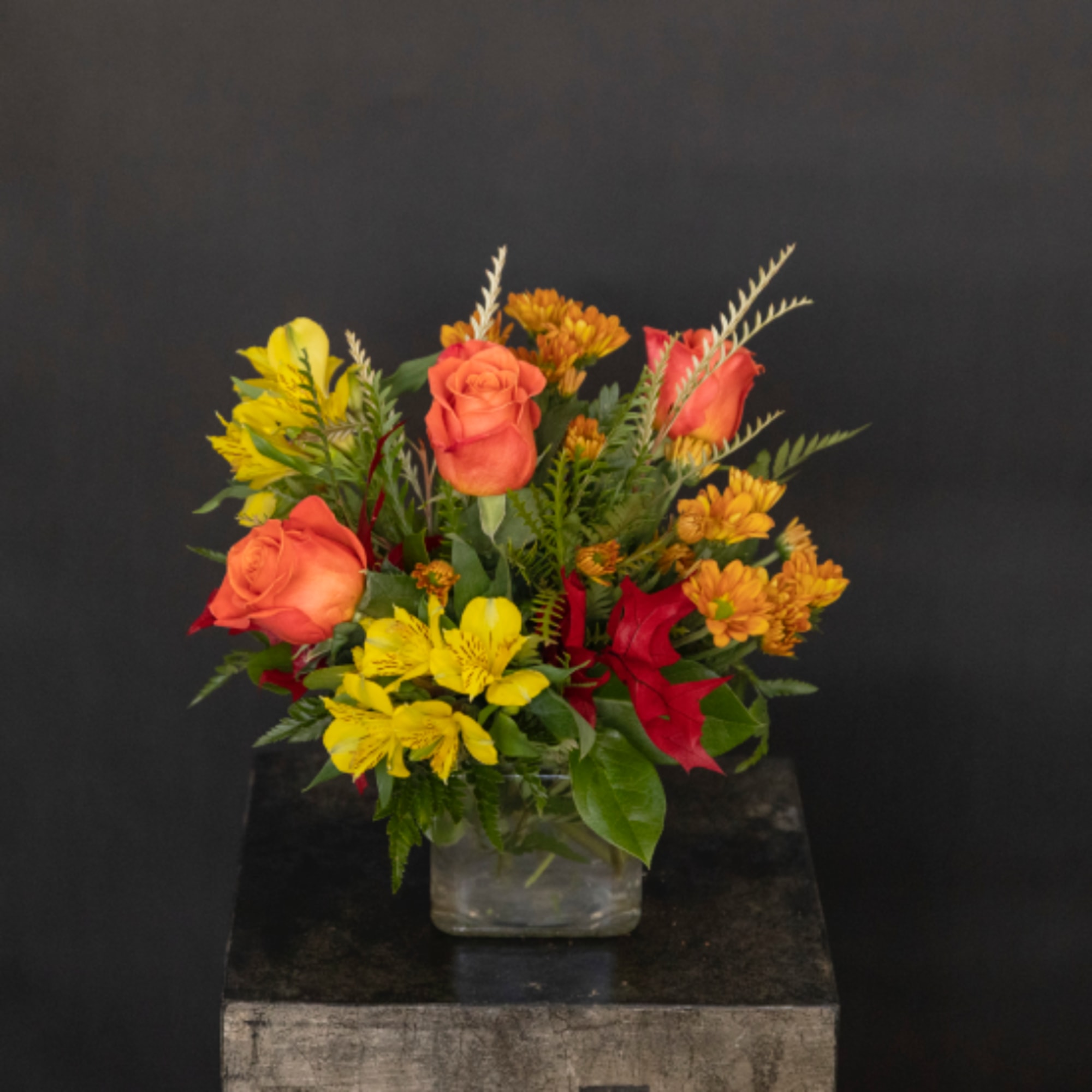 Orange and yellow roses with red flowers in a clear square vase