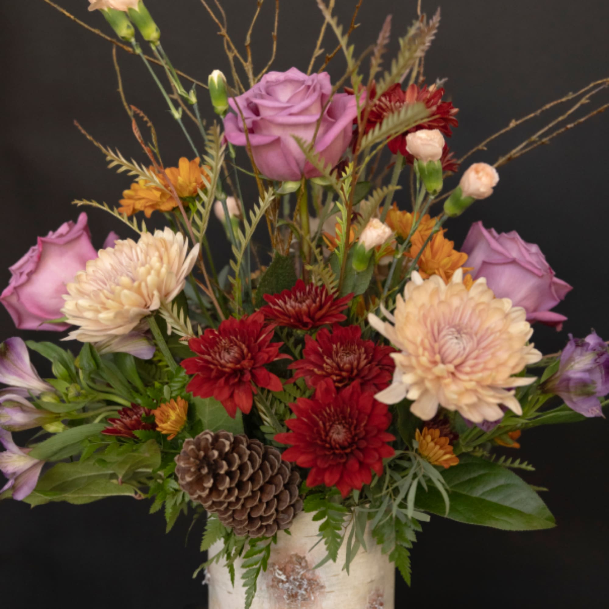 Mixed bouquet with purple roses, red and cream chrysanthemums, and a pinecone in a vase