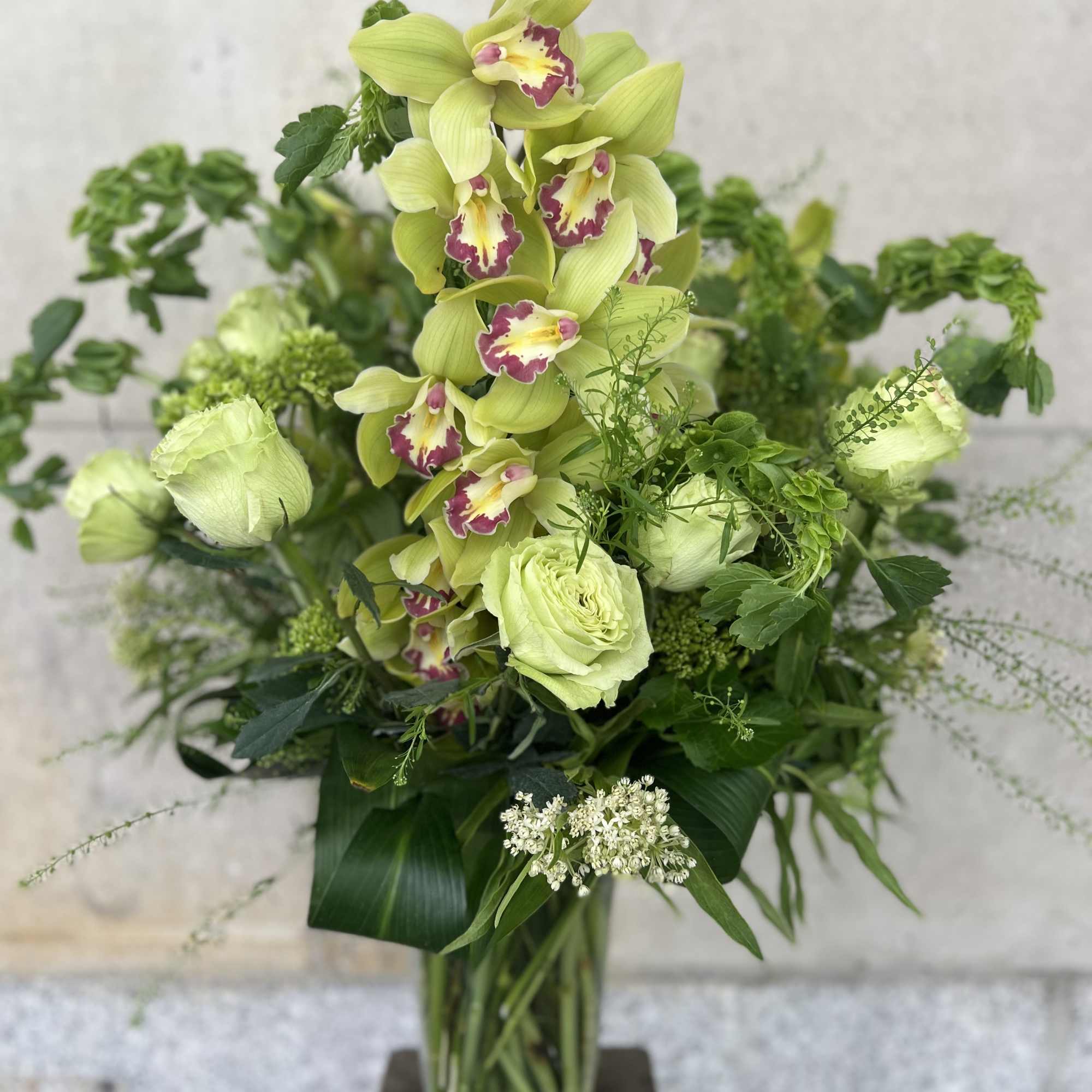 Tall green orchid arrangement in a glass vase with mixed foliage