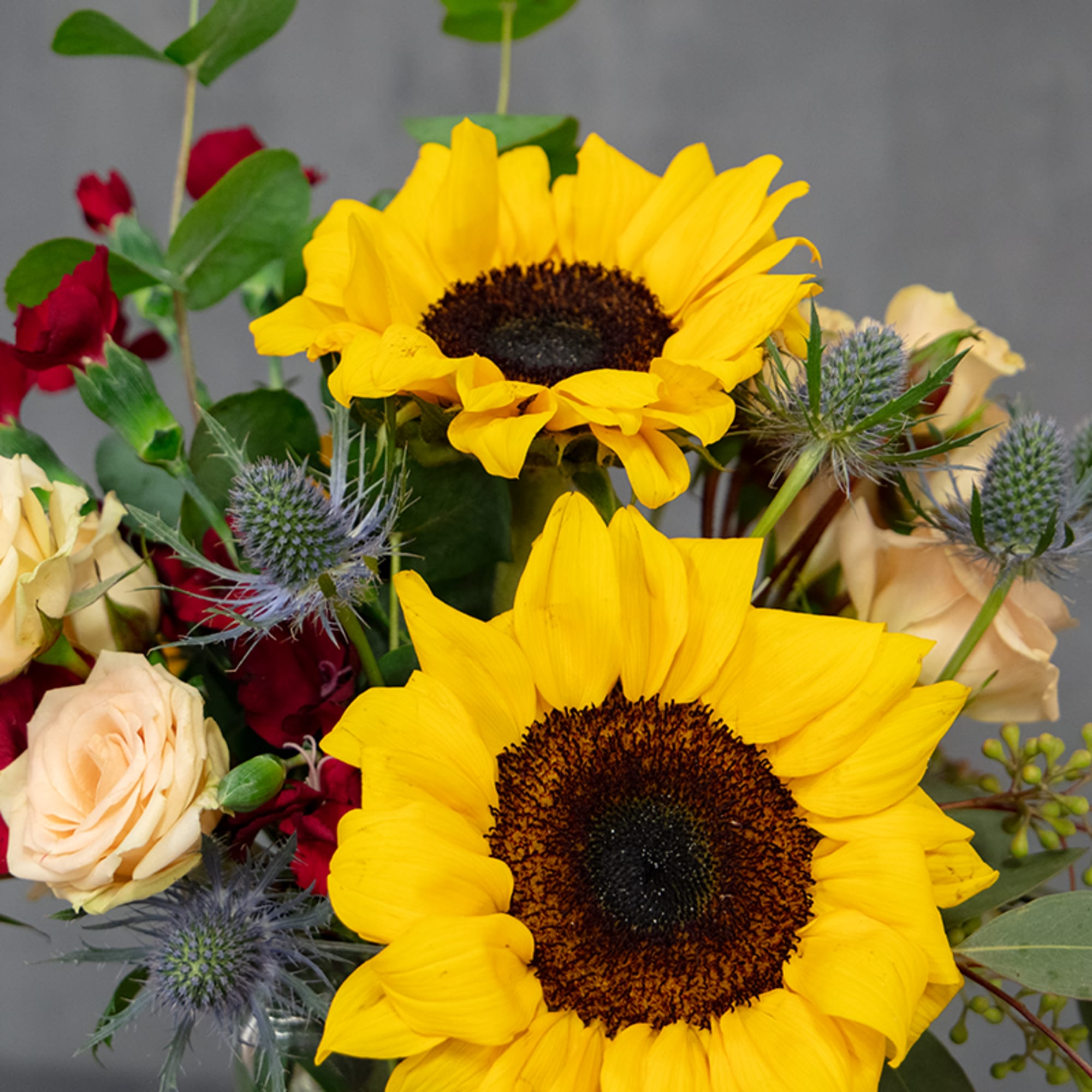 Bouquet with sunflowers, roses, and blue thistle in a glass vase