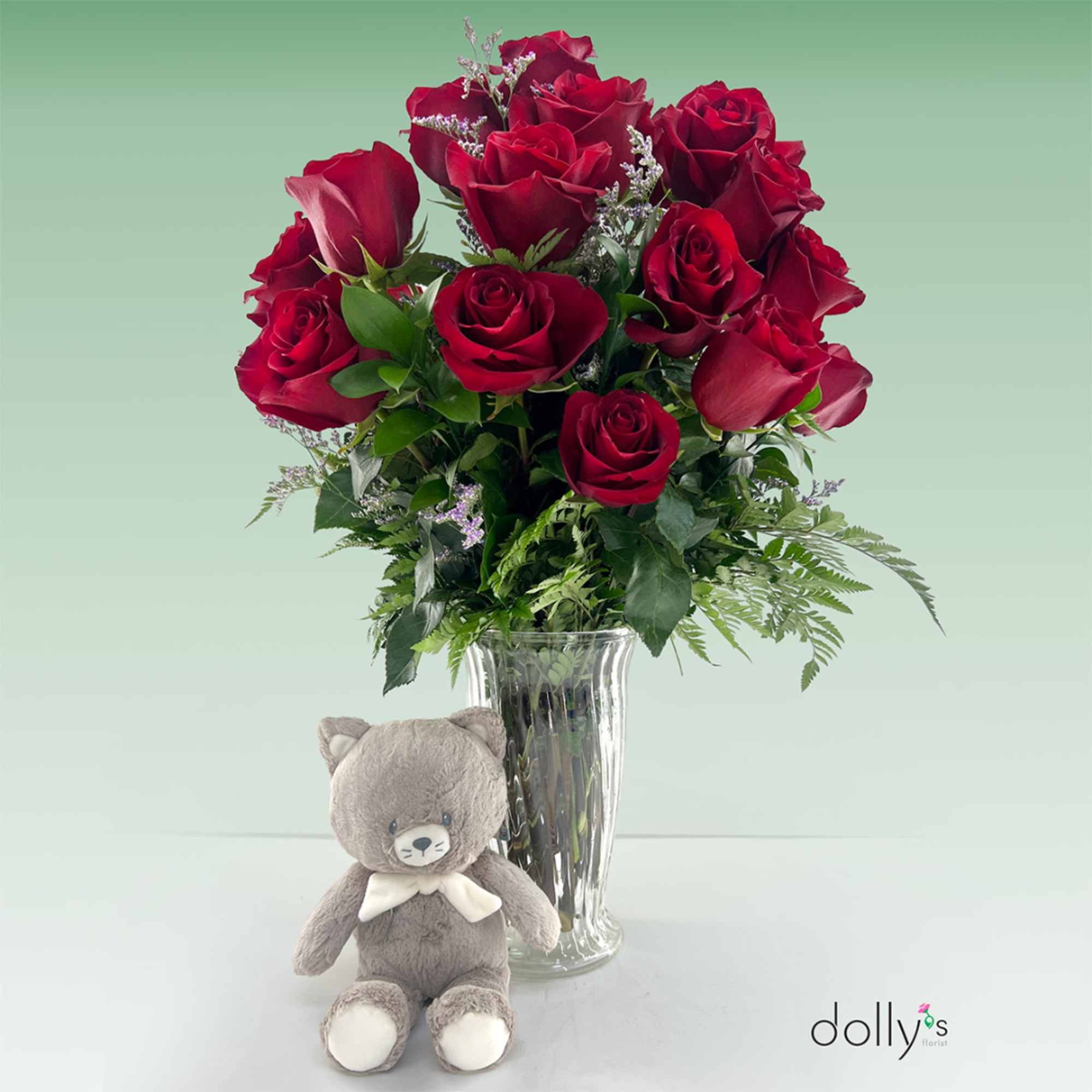 Red roses in a clear vase with a small teddy bear in front.