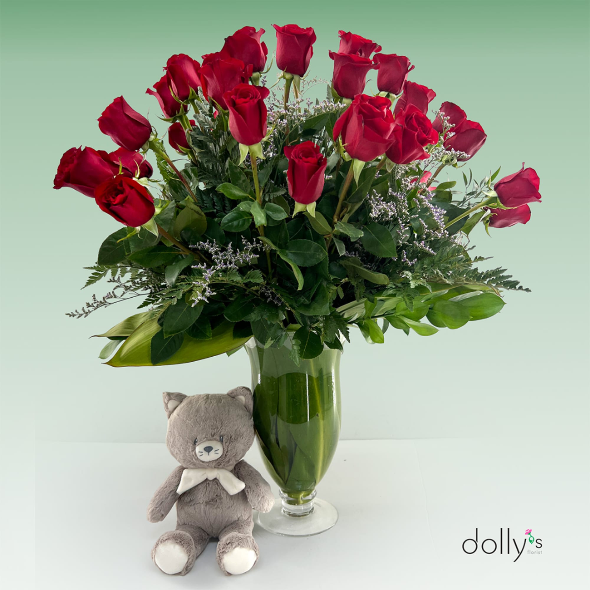 Red roses in a glass vase with a small teddy bear beside it.