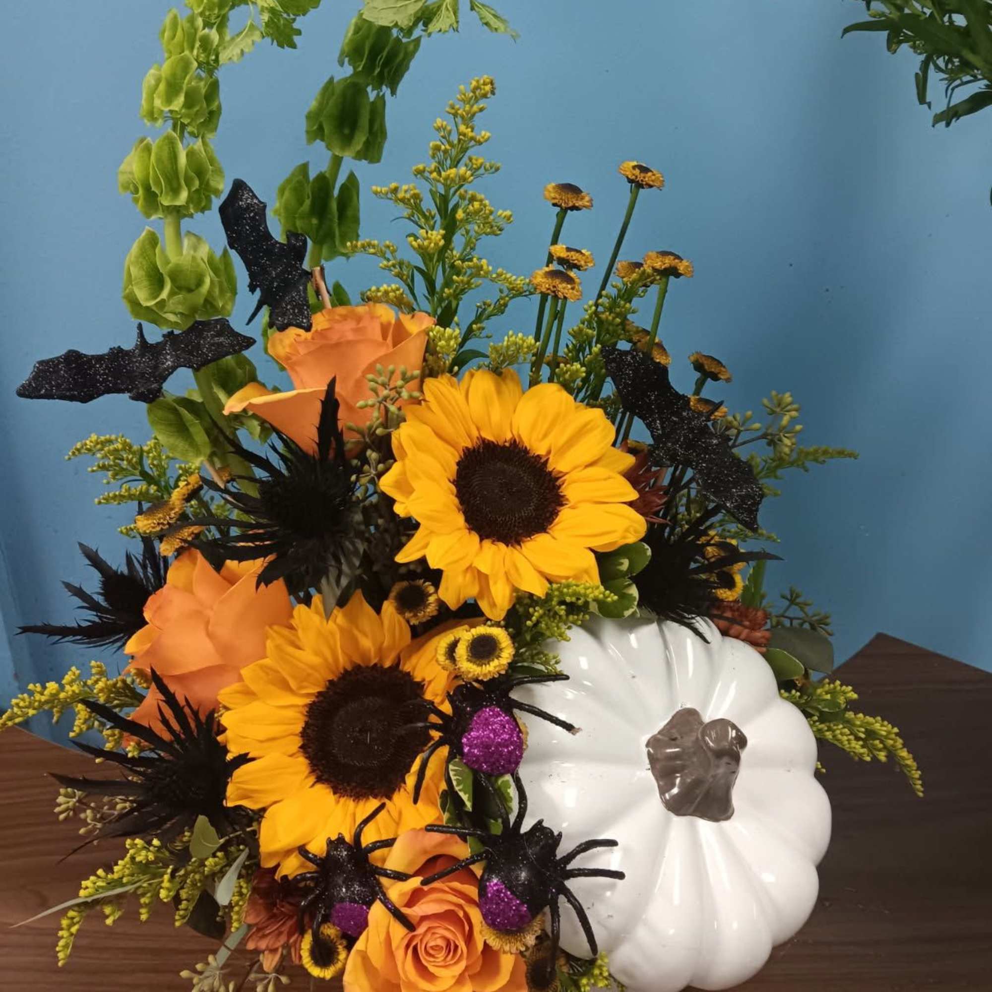 Orange roses and sunflowers arranged with Halloween spider picks in a white pumpkin container