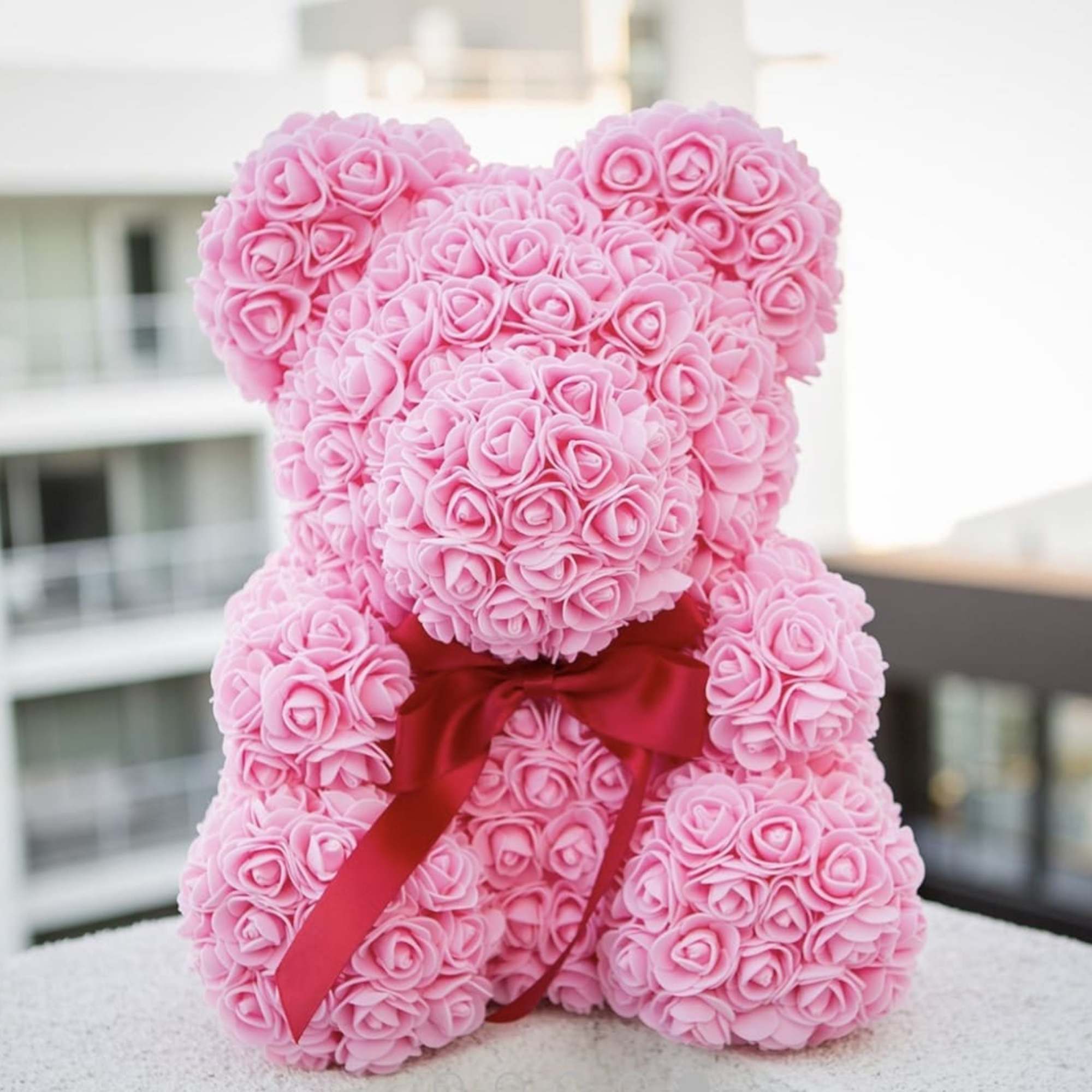Pink rose-covered teddy bear with a red ribbon bow made of small artificial rosebuds
