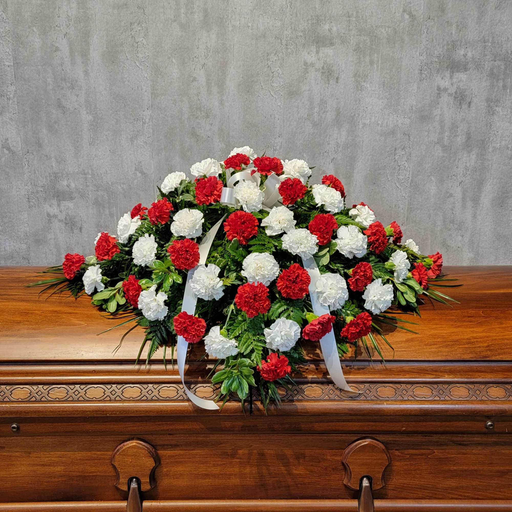 Red and white carnation casket spray with white ribbon on a wooden casket