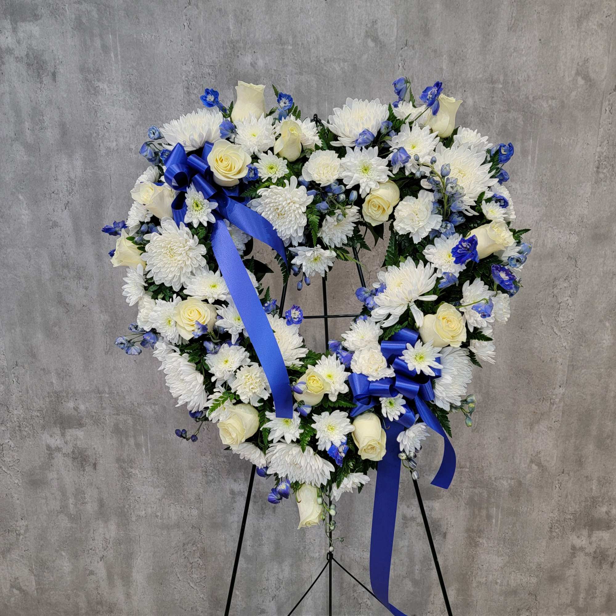 Heart-shaped floral wreath with white flowers, cream roses, and blue ribbons on a stand