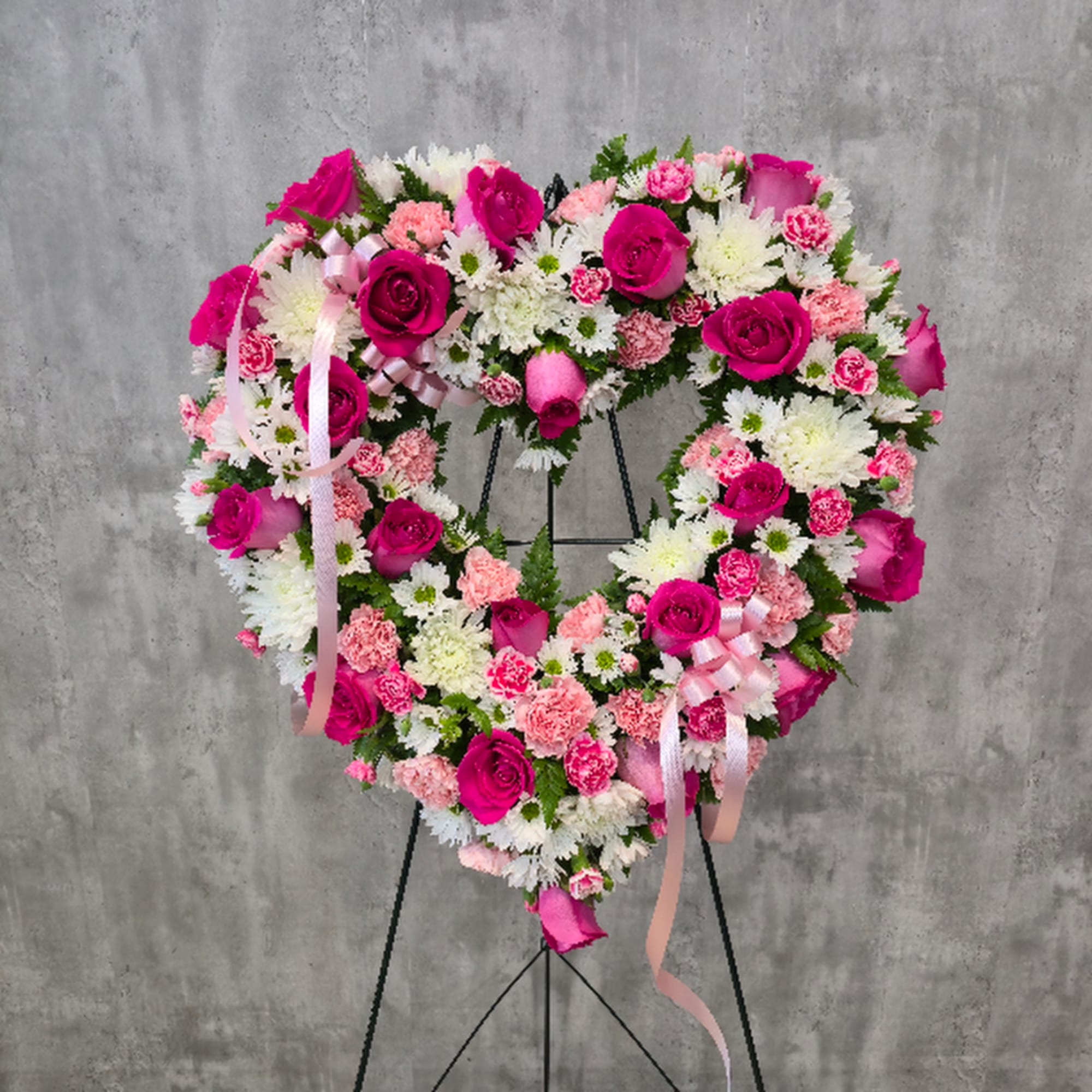 Heart-shaped floral wreath with pink roses, carnations, and white daisies on a stand