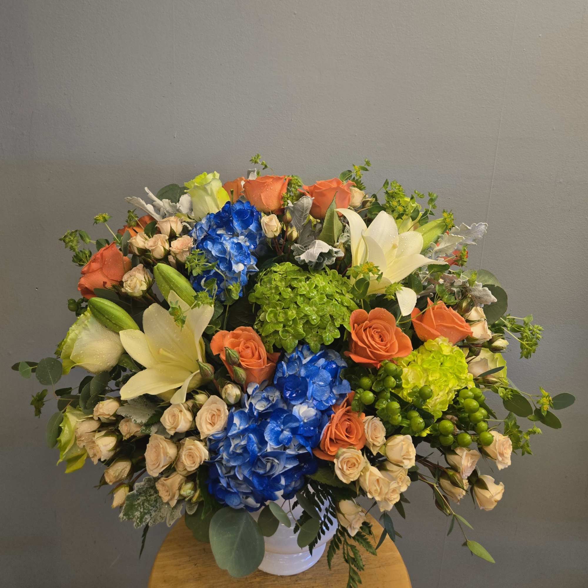 Large mixed bouquet in a white vase with orange roses, blue hydrangeas, white lilies, and green blooms