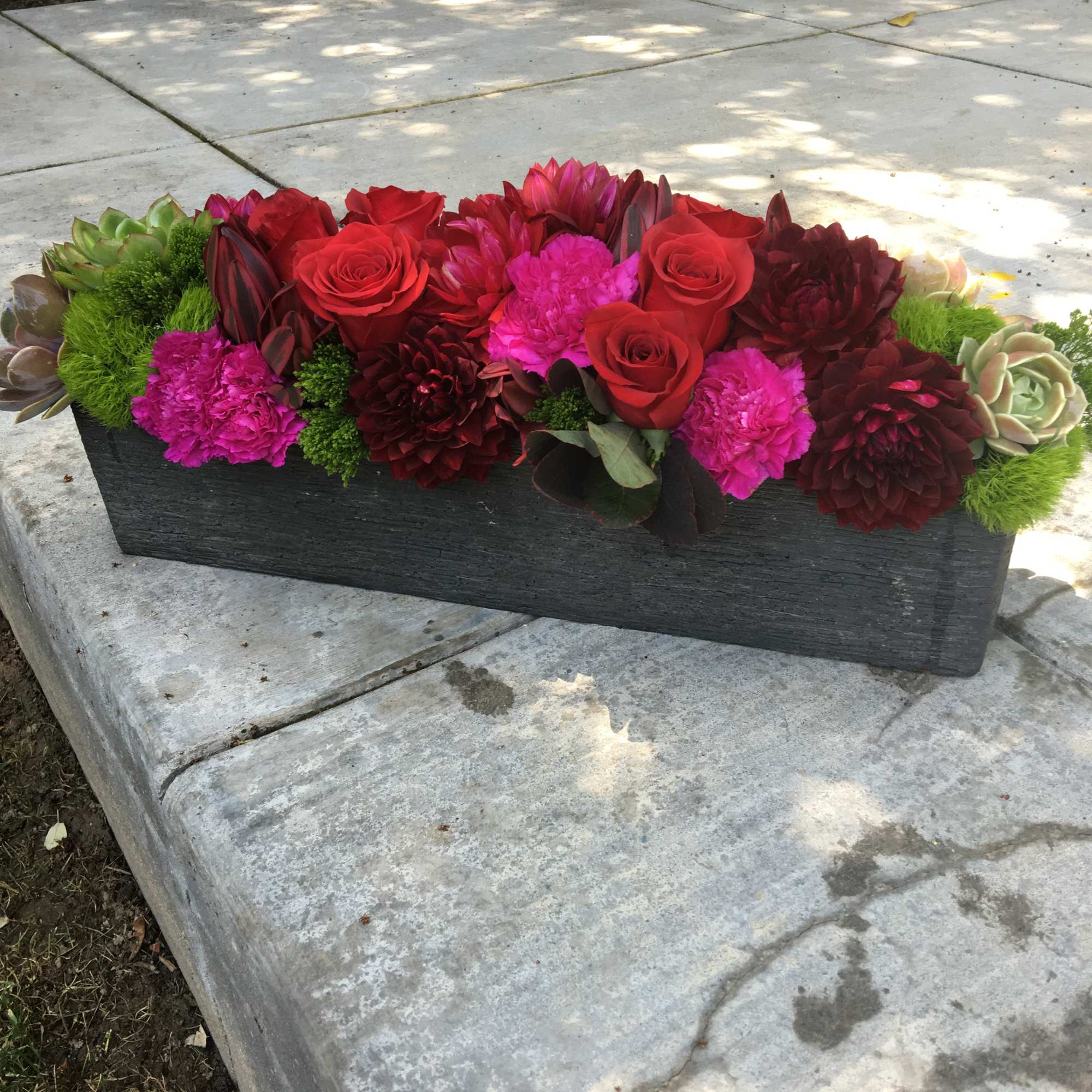 Rectangular floral arrangement with red roses, pink blooms, and succulents in a black container