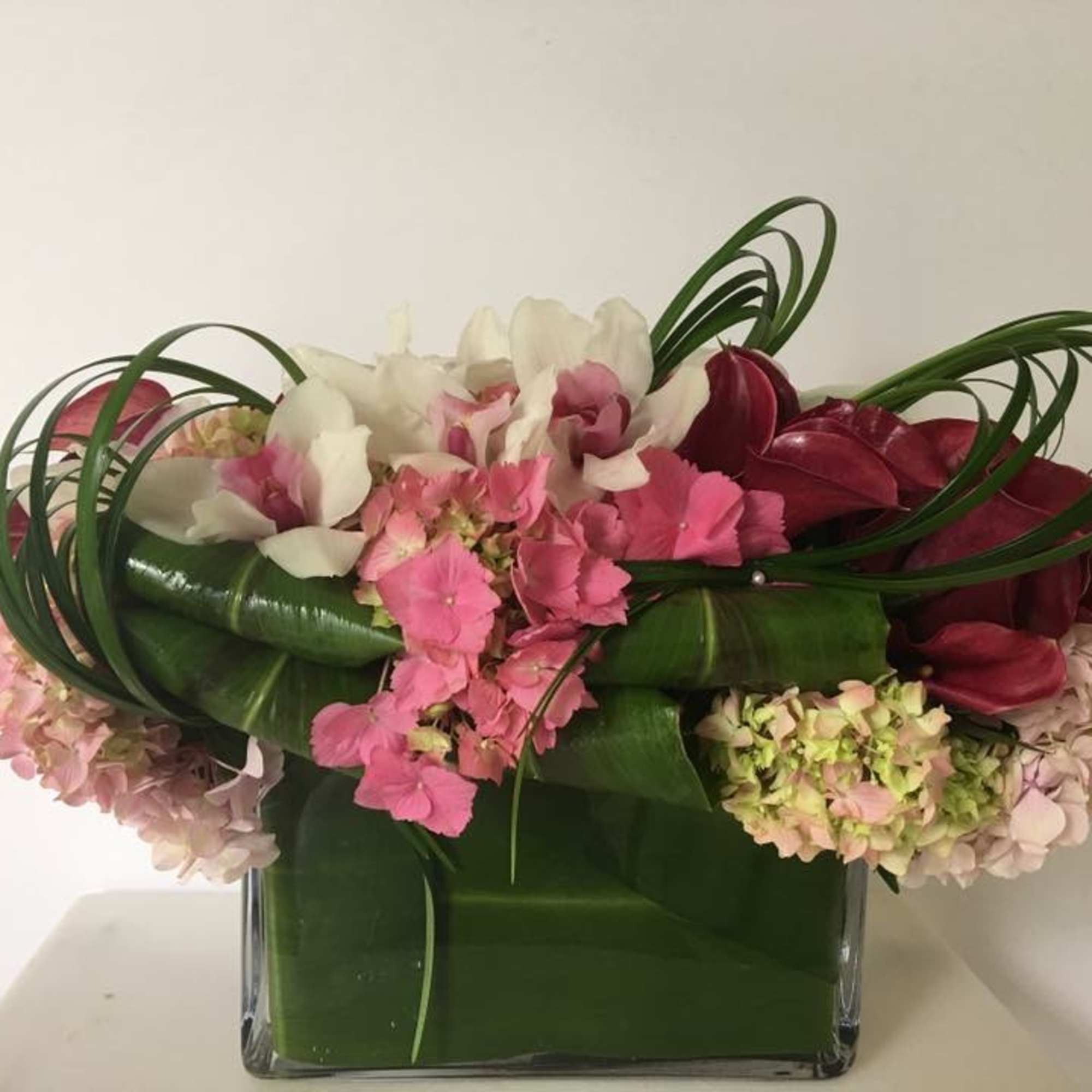 Pink and white flowers arranged in a square glass vase with looping green leaves