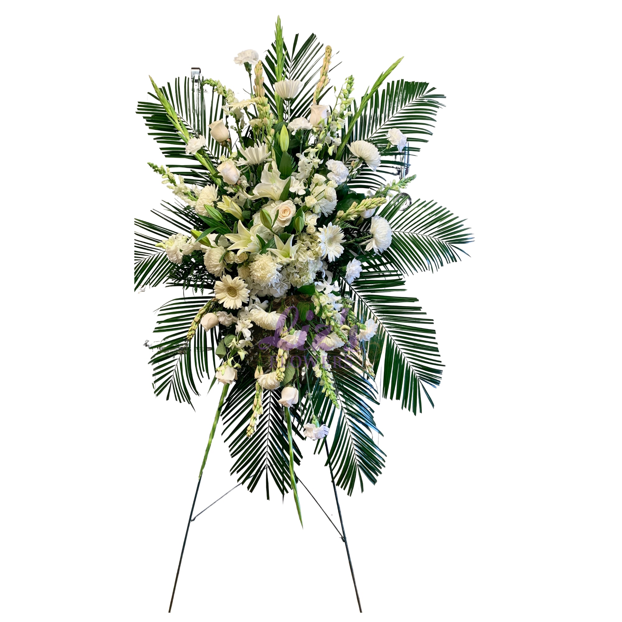 Tall white standing spray with roses, lilies, and other flowers framed by large palm fronds on an easel.