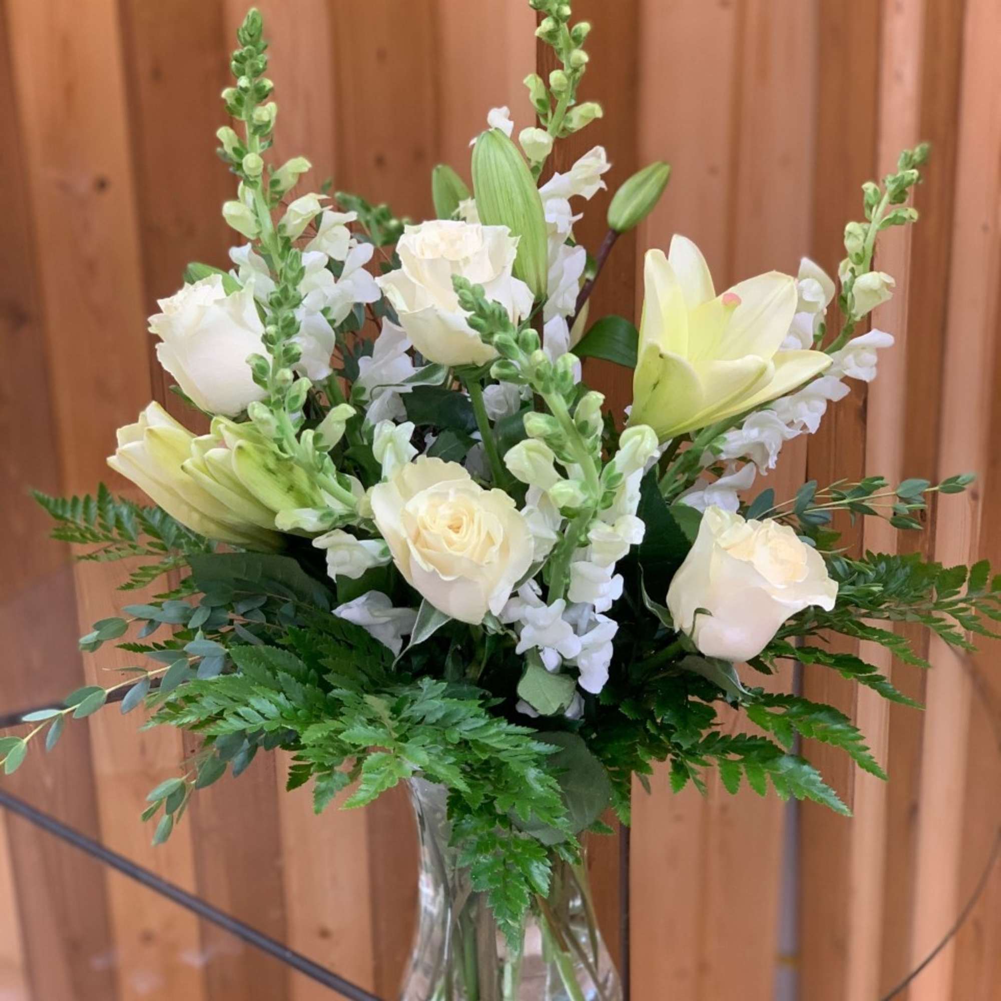 White roses, lilies, and snapdragons arranged in a clear glass vase with lush foliage.