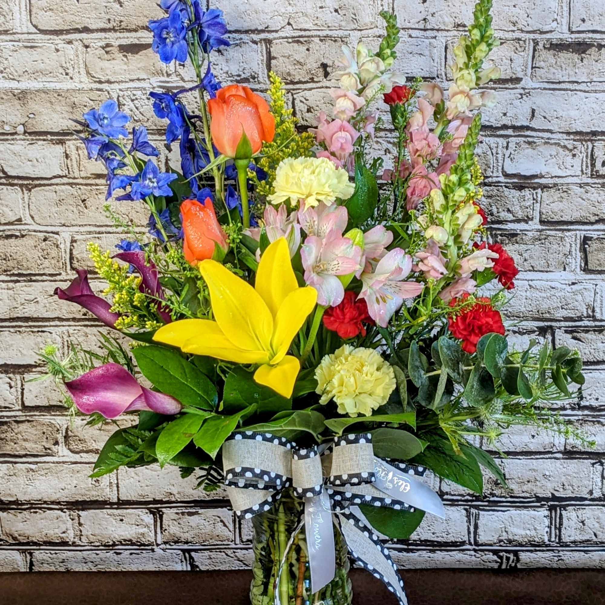 Tall mixed-color flower arrangement in a clear glass vase with a ribbon bow.