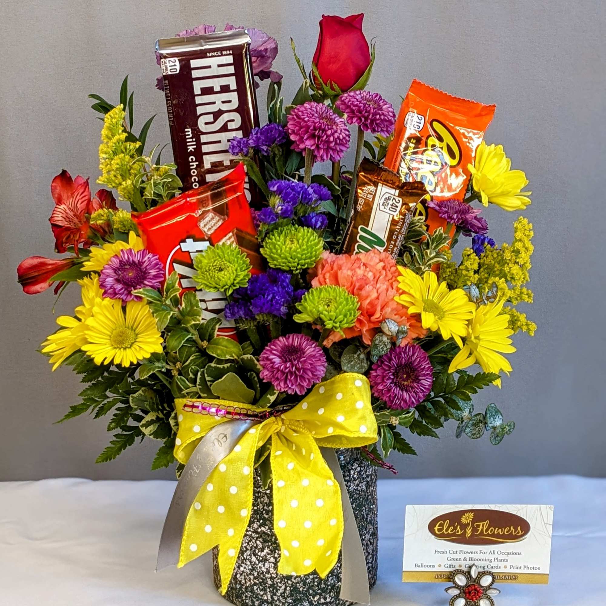 Bright mixed flower arrangement with candy bars in a speckled container tied with a yellow polka-dot bow.
