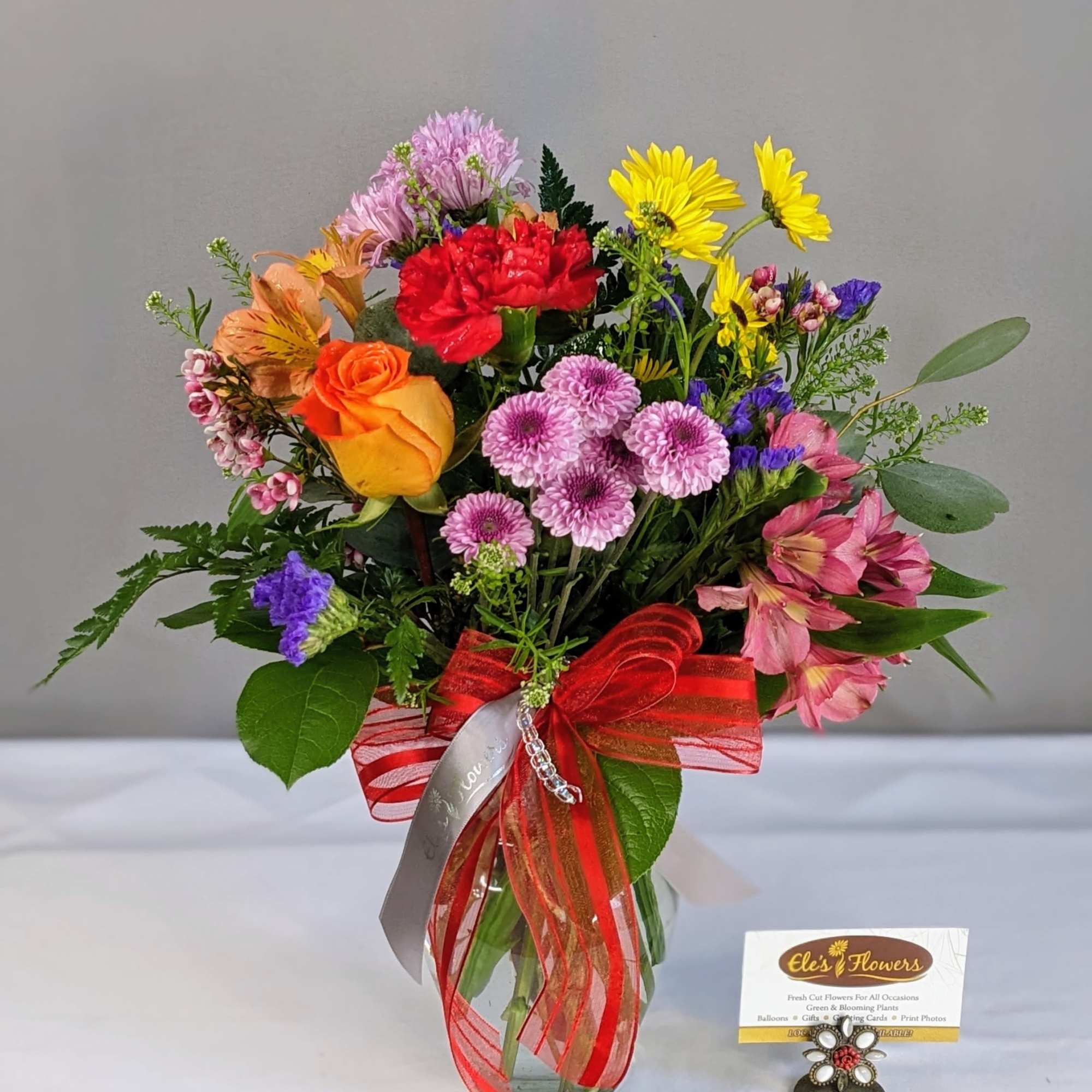 Bright mixed bouquet of roses, carnations, daisies, and mums in a clear vase with a red bow.