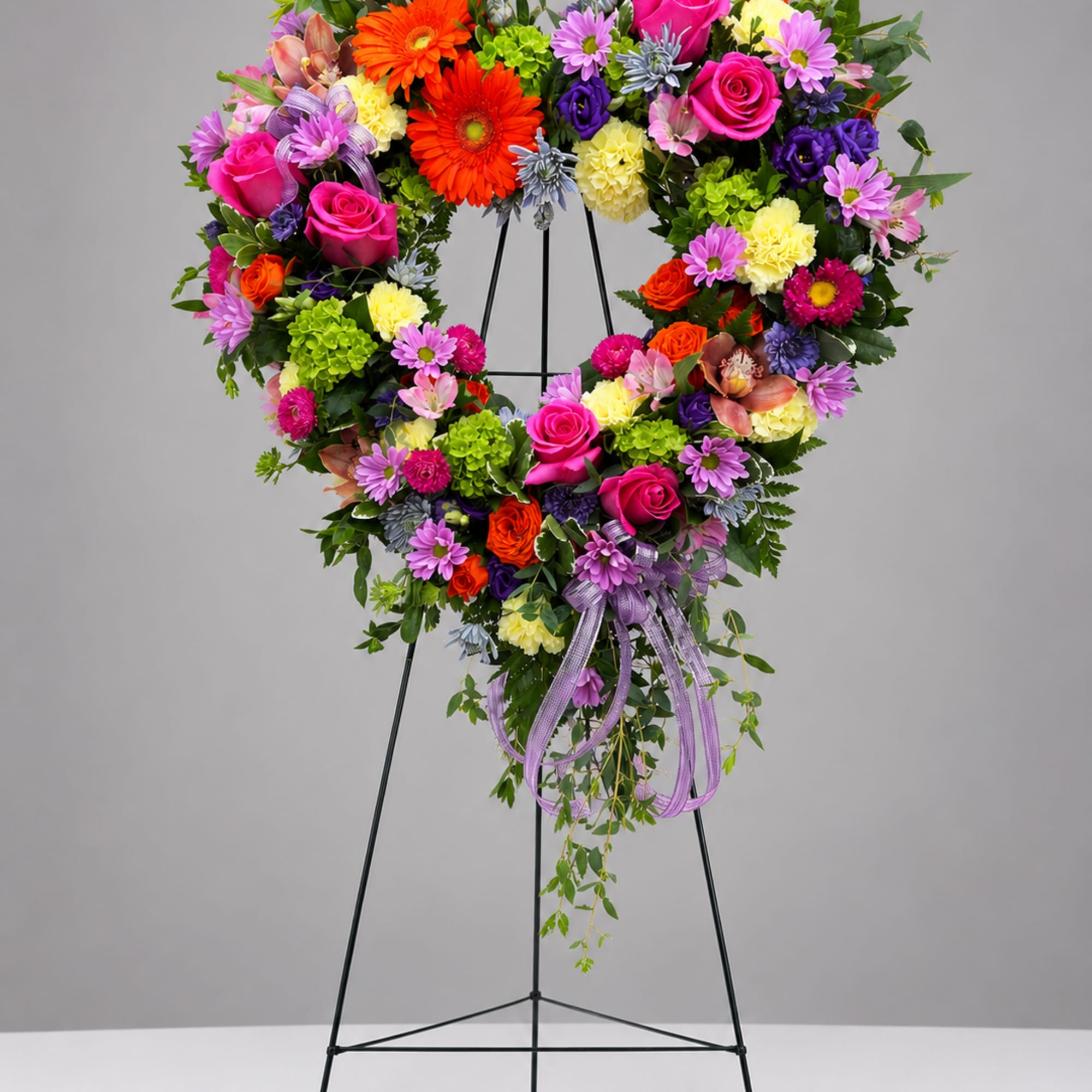 Heart-shaped floral wreath on a stand with bright mixed blooms and a purple ribbon