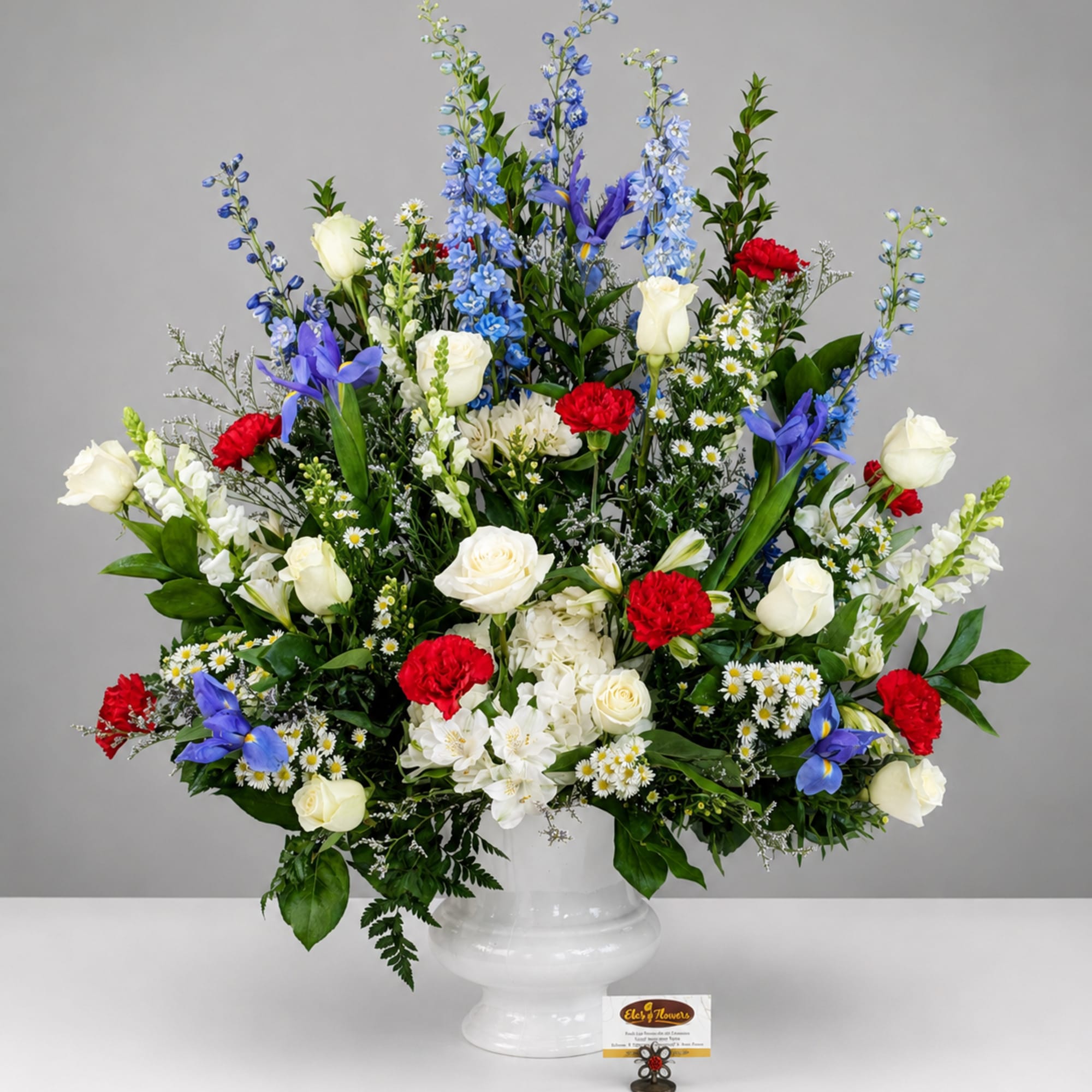 Large mixed flower arrangement in a white urn vase with blue, white, and red blooms.