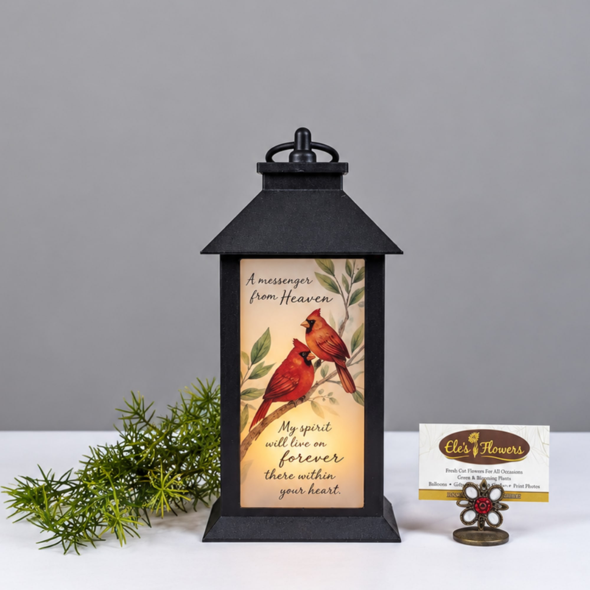 Black lantern with a cardinal memorial insert and a small greenery sprig beside it