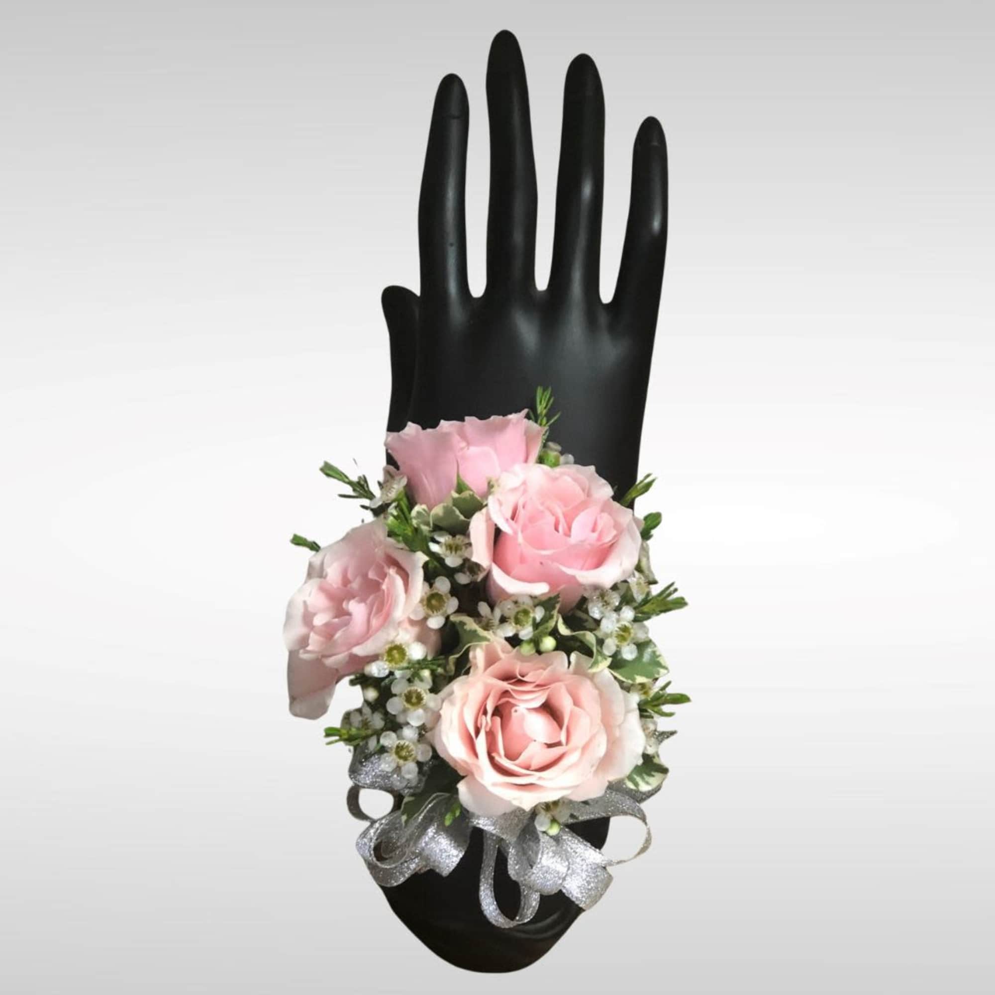 Pink rose wrist corsage with small white blooms and silver ribbon on a black hand display.