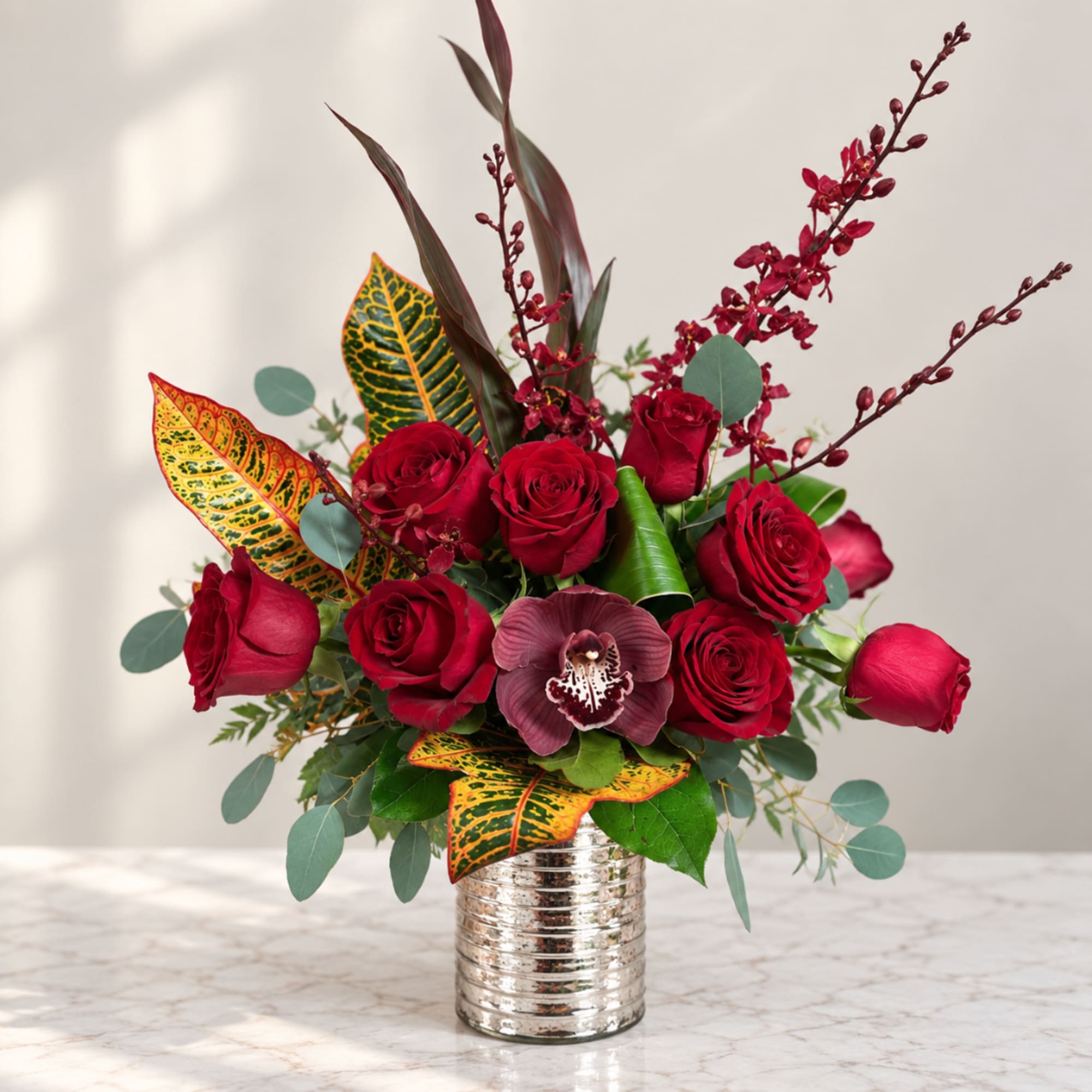 Red roses arranged in a silver vase with burgundy orchids and patterned leaves