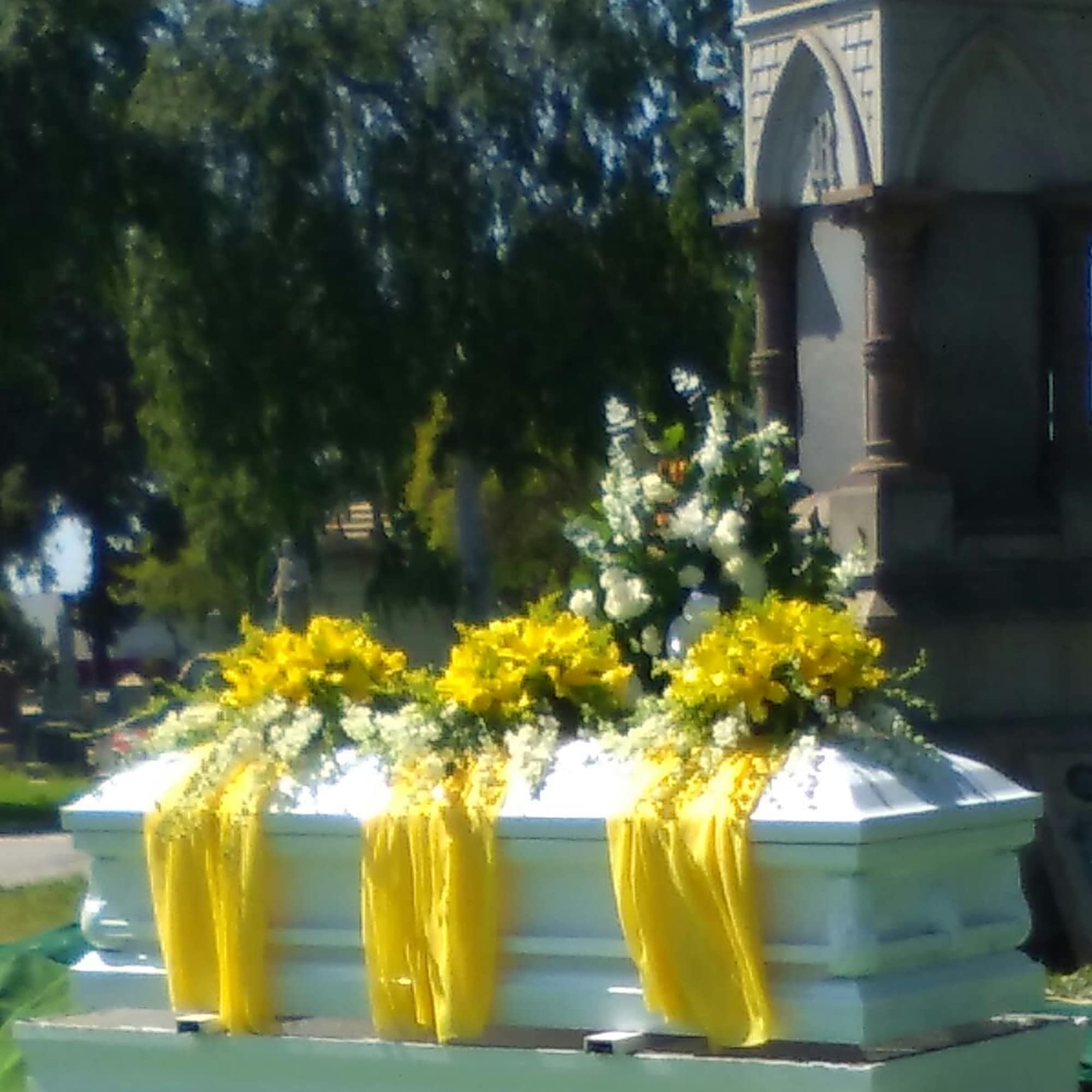 White casket with yellow floral sprays and draped yellow fabric outdoors