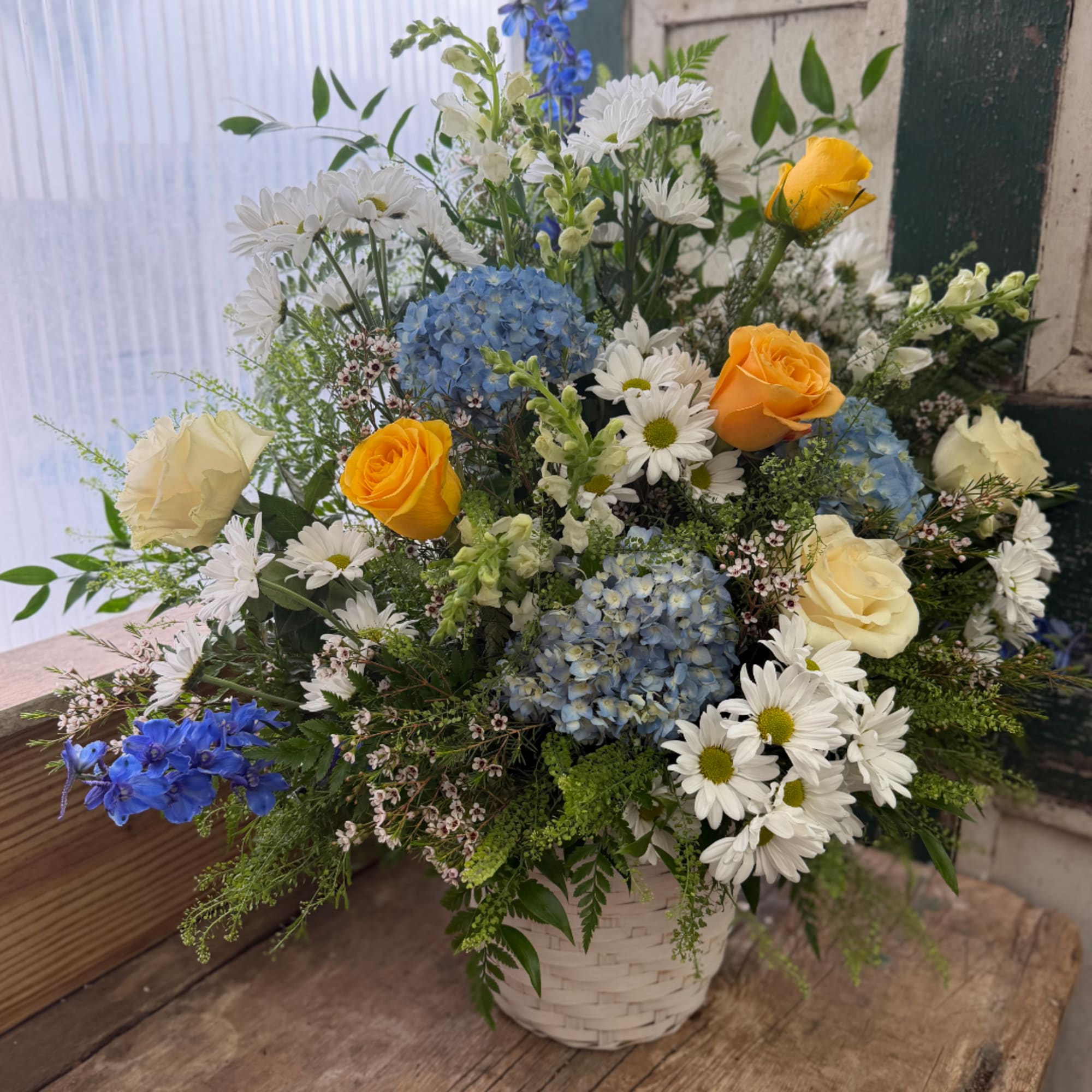 Mixed bouquet of blue hydrangeas, white daisies, and yellow roses in a basket