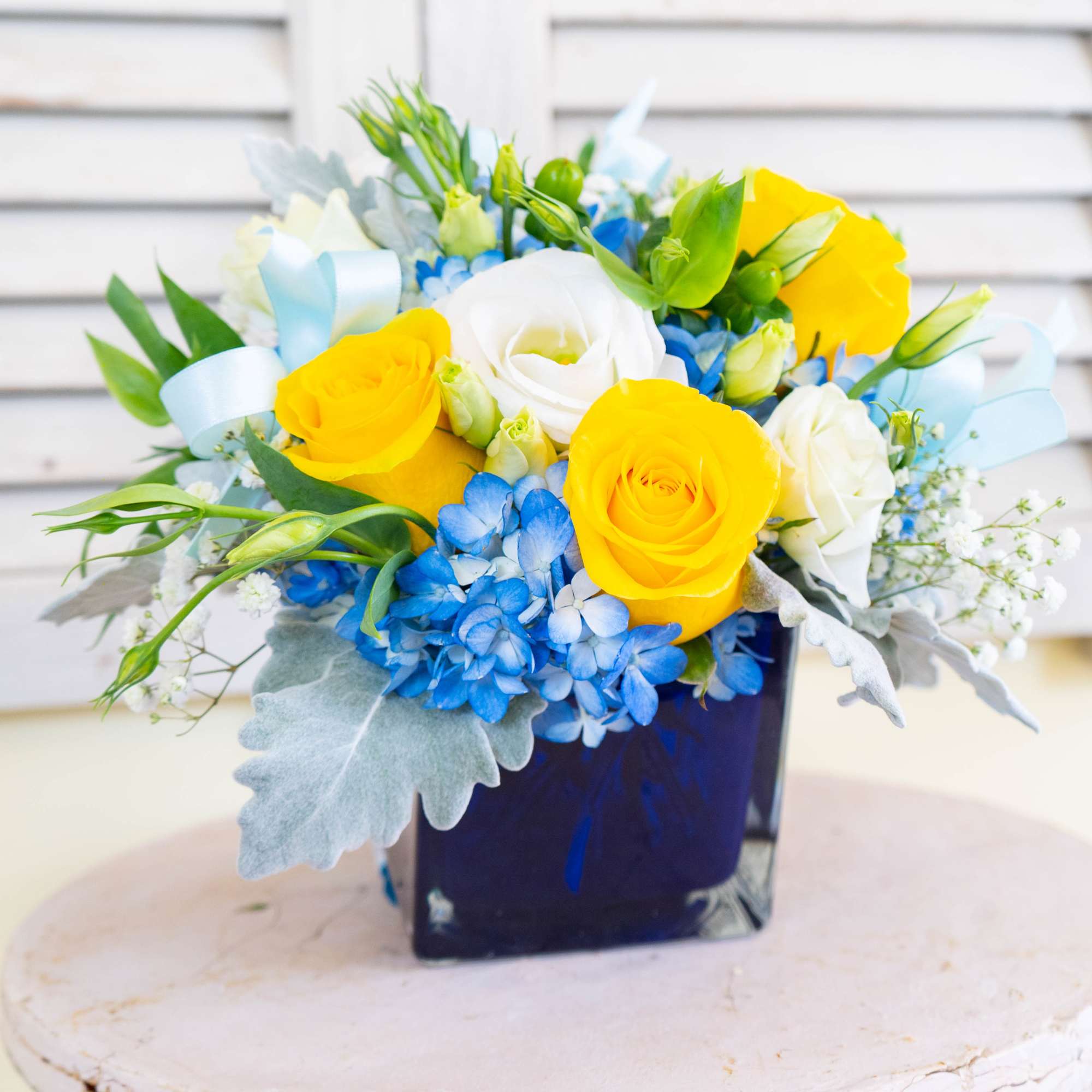 Yellow and white roses with blue hydrangeas in a dark square vase