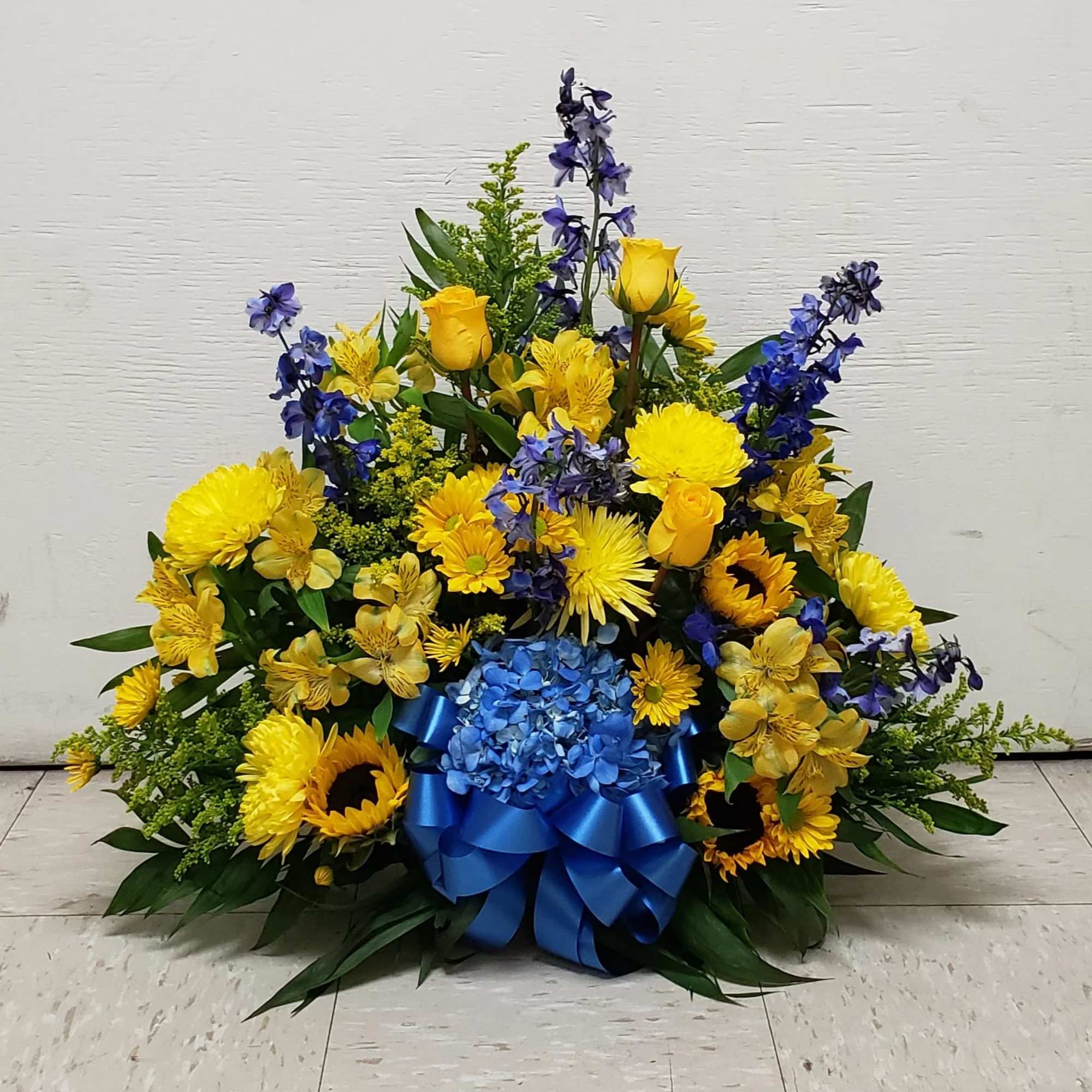 Fan-shaped yellow and blue arrangement with sunflowers, roses, delphinium, and a blue ribbon bow.
